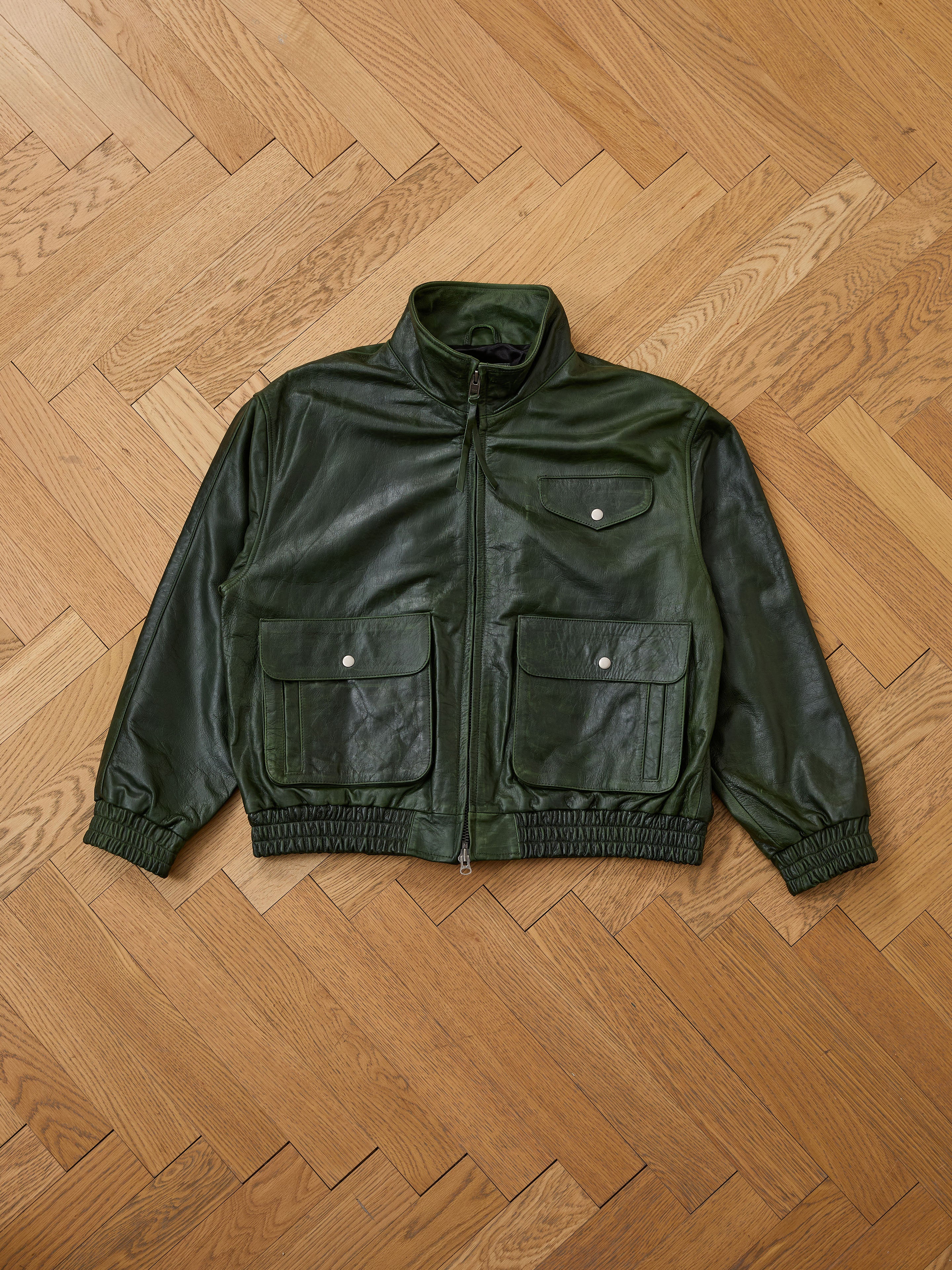The FOUND Forest Leather Bomber Jacket in green, featuring three front pockets and elastic cuffs, is shown laid flat on a parquet wooden floor.