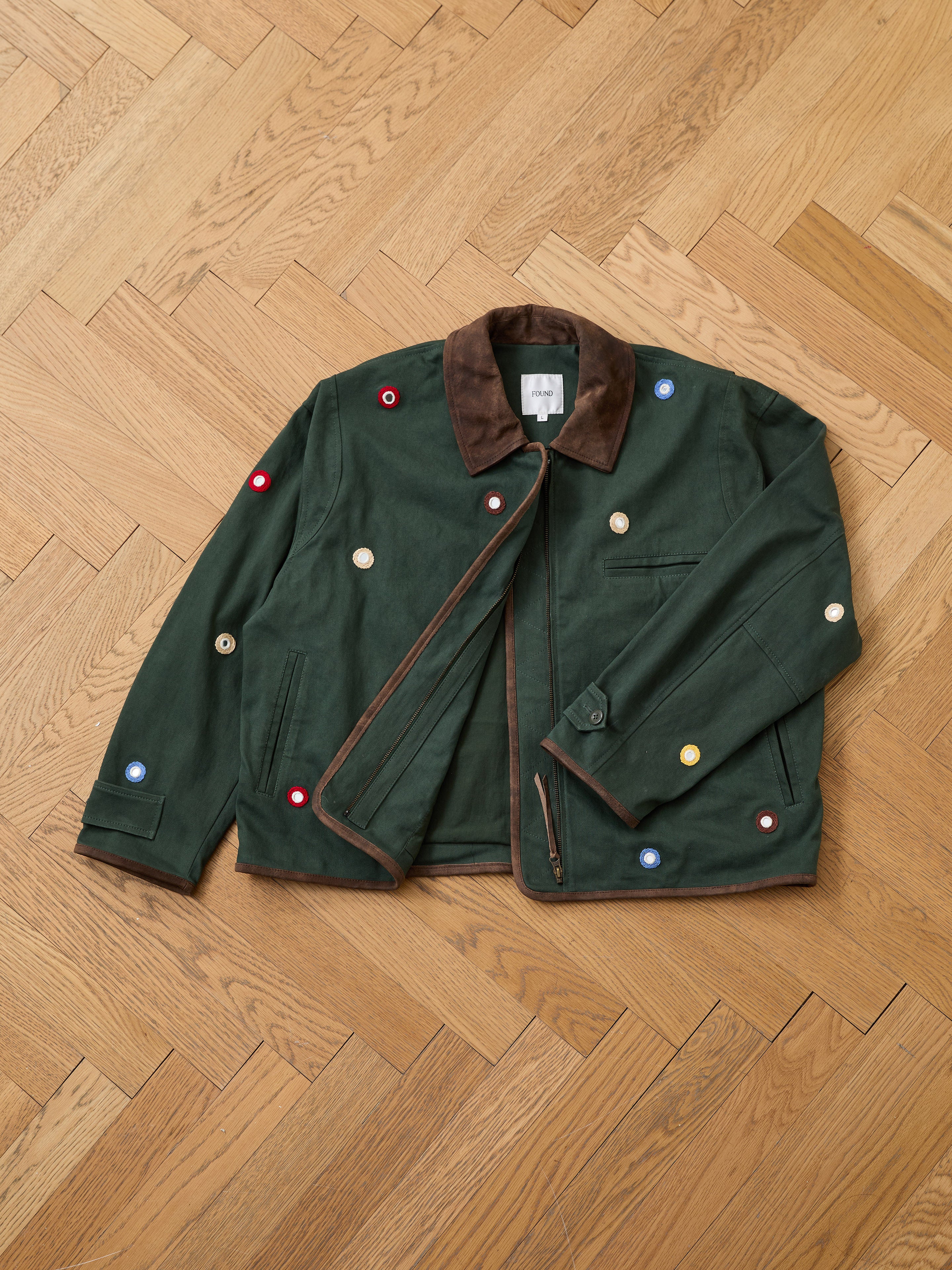 The Hand Embroidered Mirror Abhala Jacket by Found, featuring a brown collar and multicolored circular patches, is laid flat on a wooden parquet floor.