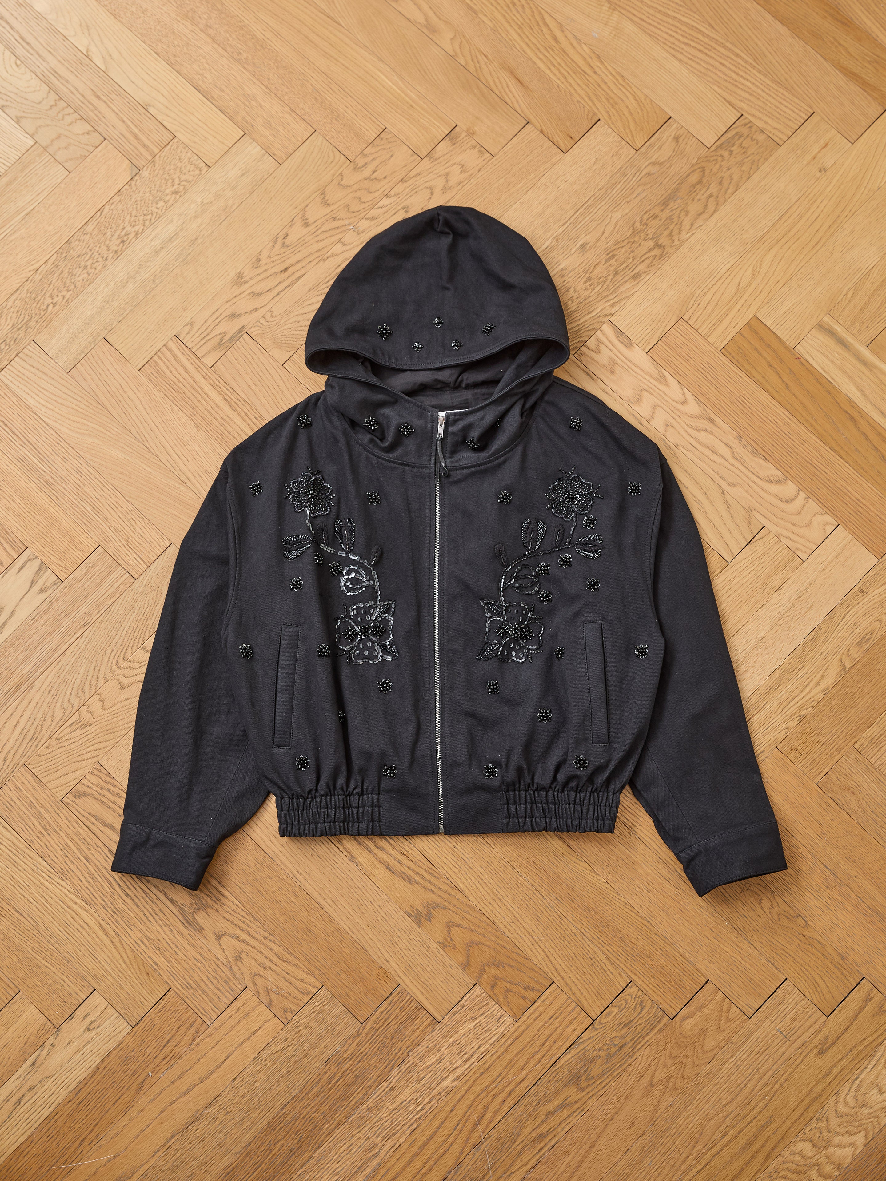 The FOUND Hand Embroidered Tonal Anorak Jacket is a black unisex cotton piece with Salma Sitara floral hand-embroidery and bead embellishments, shown flat on a wooden herringbone floor.