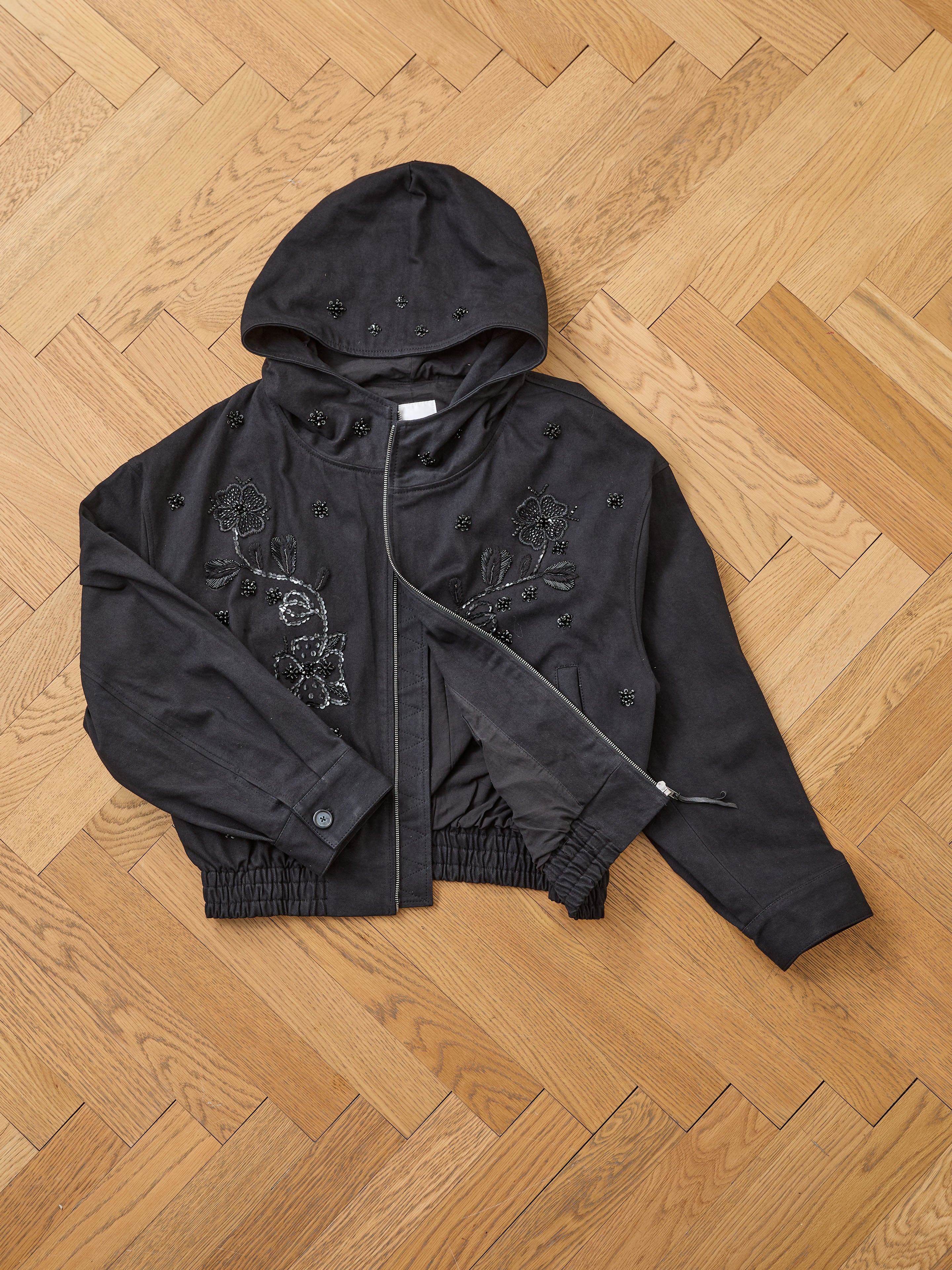 The FOUND Hand Embroidered Tonal Anorak Jacket, a black unisex cotton piece with Salma Sitara embroidery and a partially open front zipper, is laid flat on a wooden herringbone floor.