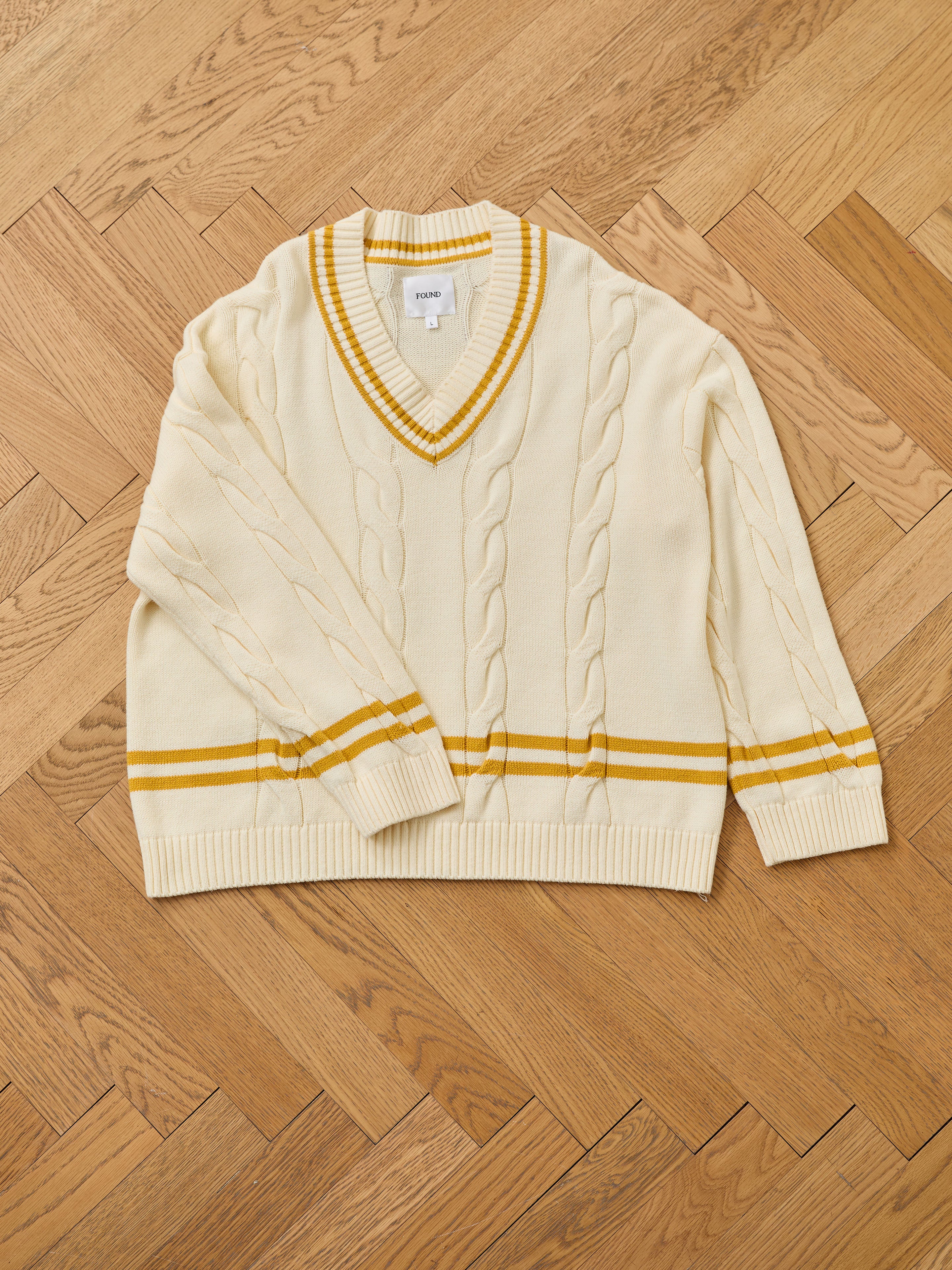 A cream-colored FOUND V-Neck Cricket Sweater with yellow stripes and classic cable knit design is laid flat on a wooden herringbone floor.