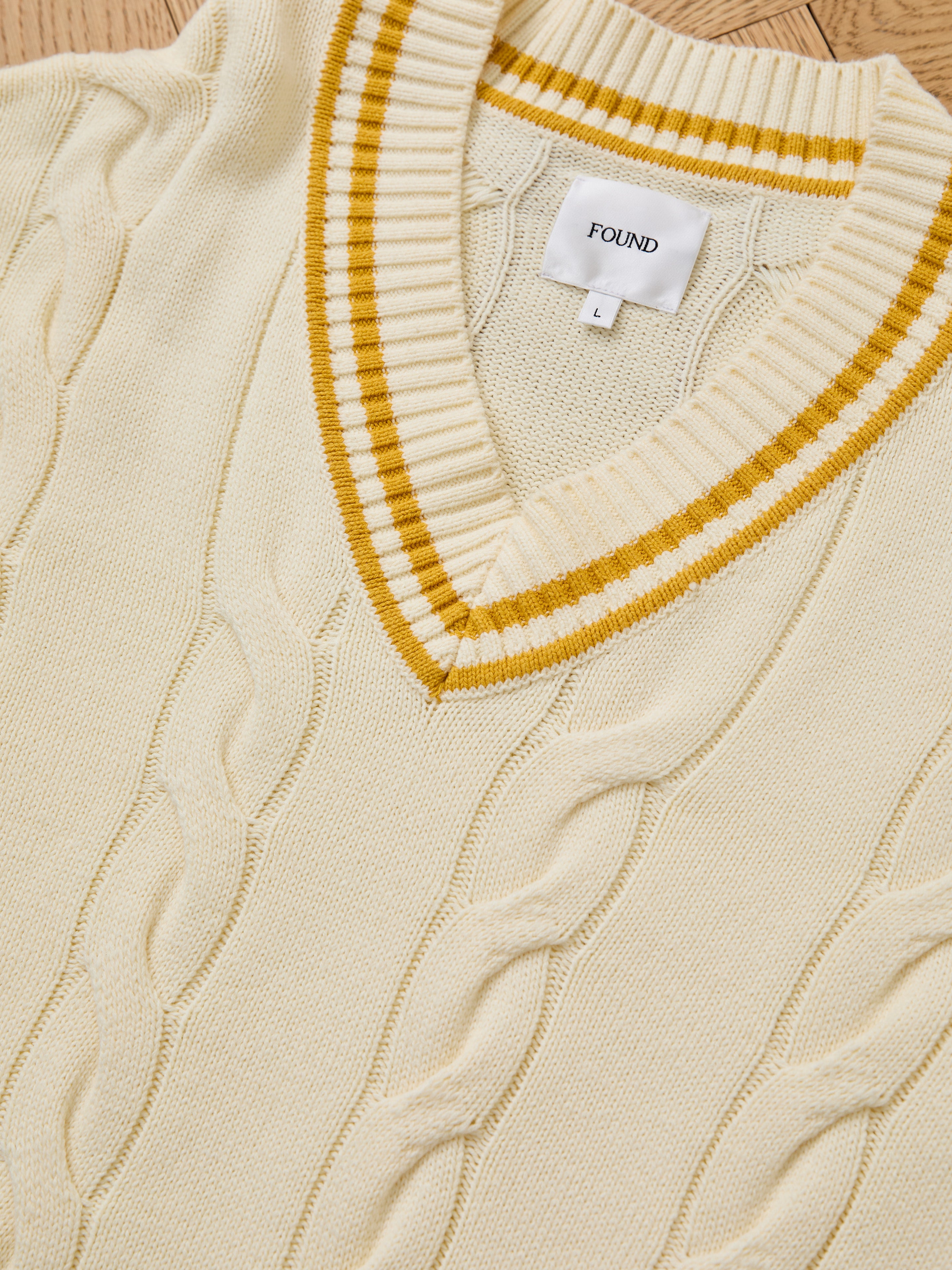 The FOUND V-Neck Cricket Sweater, in size L, features a cream cable knit with yellow stripes. This unisex sweater blends classic style and comfort and is displayed flat on a wooden surface.