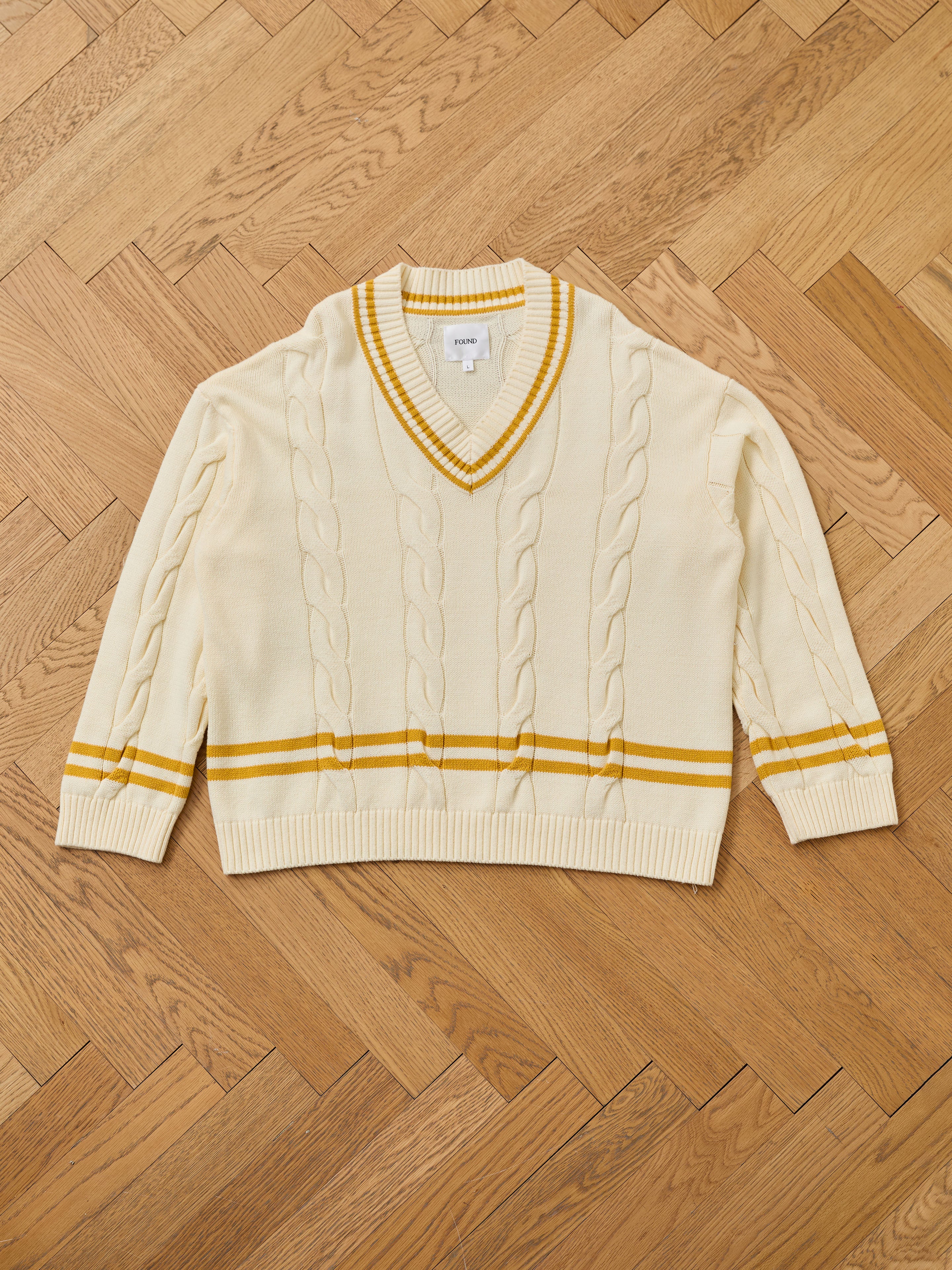 FOUND's V-Neck Cricket Sweater is a cream unisex cable knit with yellow stripes on the sleeves and hem, shown on a wooden herringbone floor.