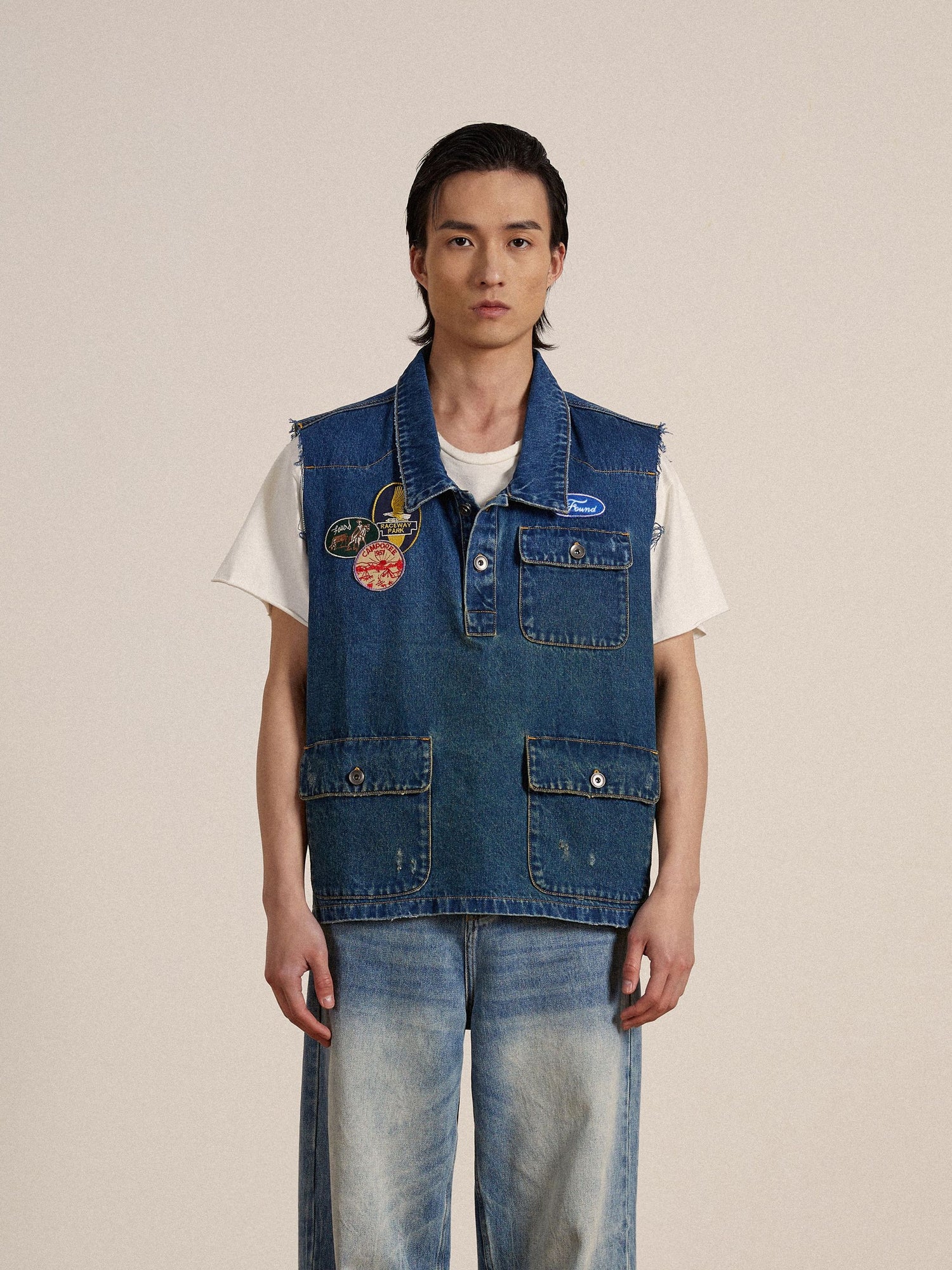 A young man stands wearing a sleeveless Found Raw Cut Patch Mechanic Denim Vest adorned with embroidered patches over a denim shirt and jeans, in front of a neutral backdrop.
