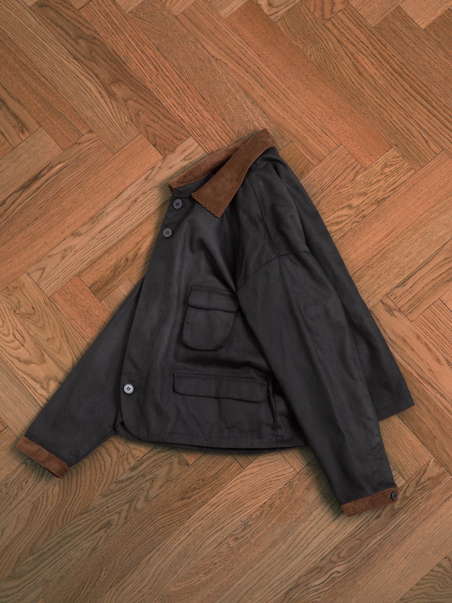 A black Lar Waxed Cotton Box Coat laying on a wooden floor by Found.