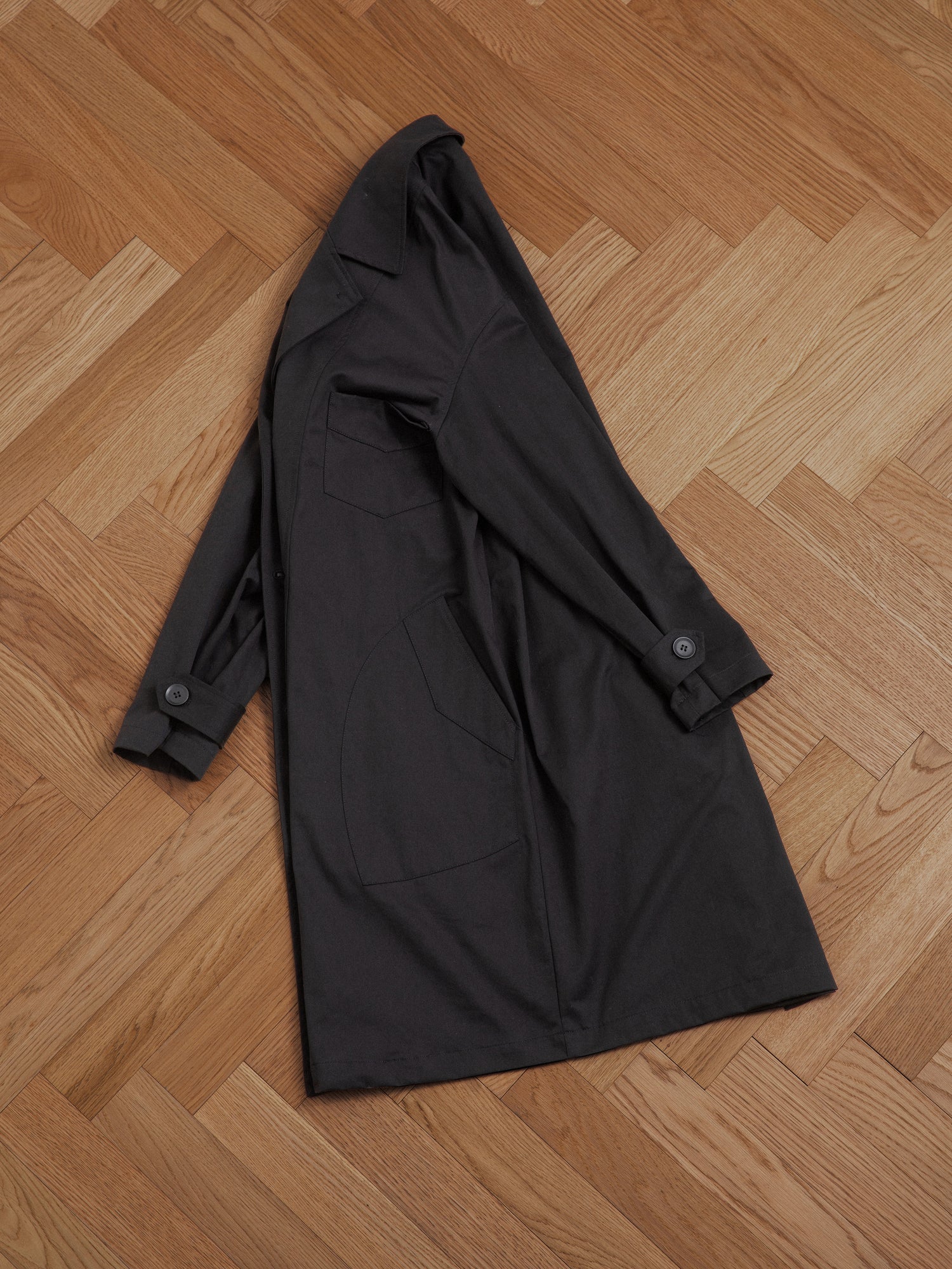 A black Found naval trench coat laying on a wooden floor, exuding enduring sophistication.