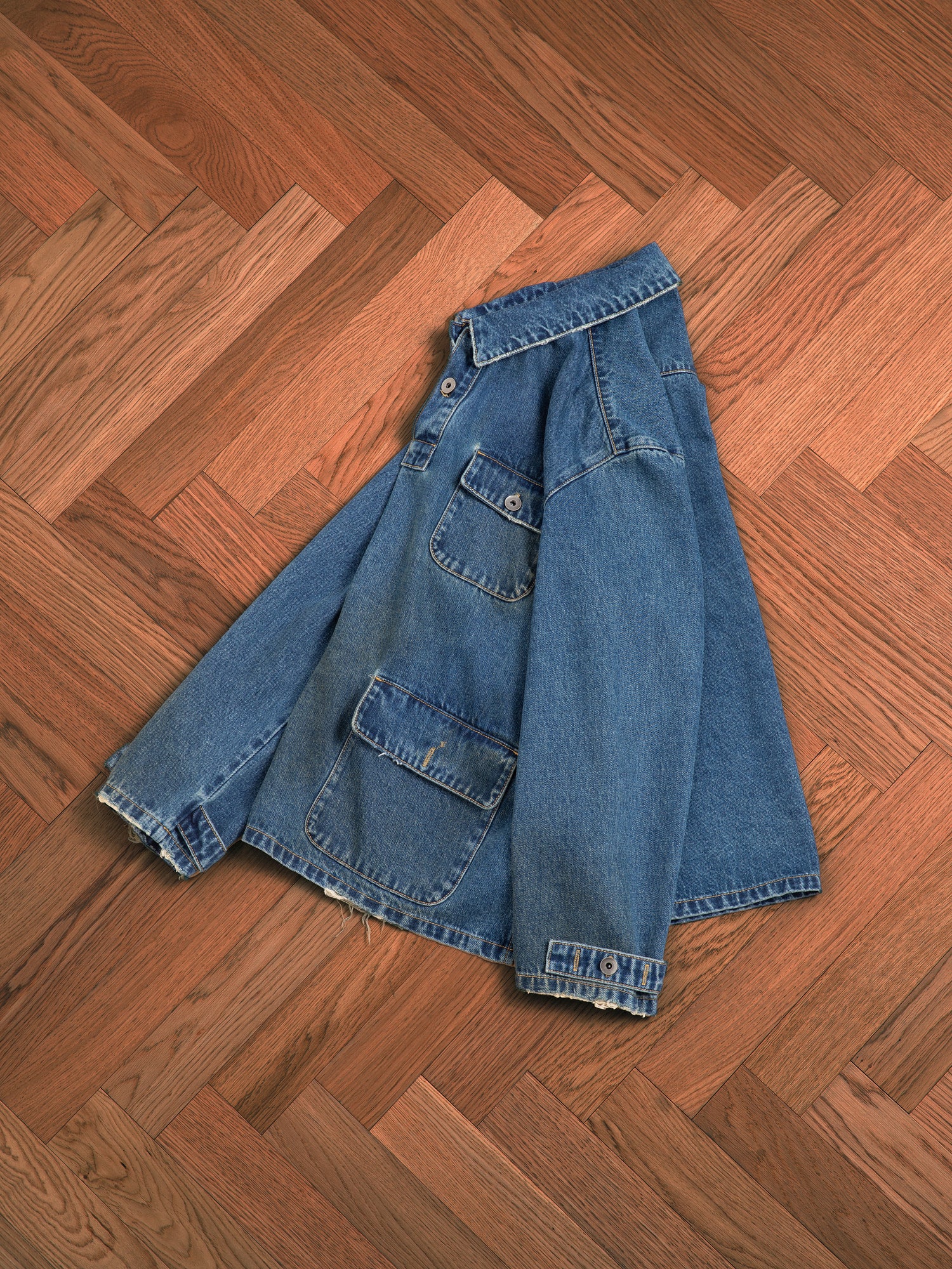 A Found blue denim jacket made from premium denim fabric, lying flat on a herringbone-patterned wooden floor.