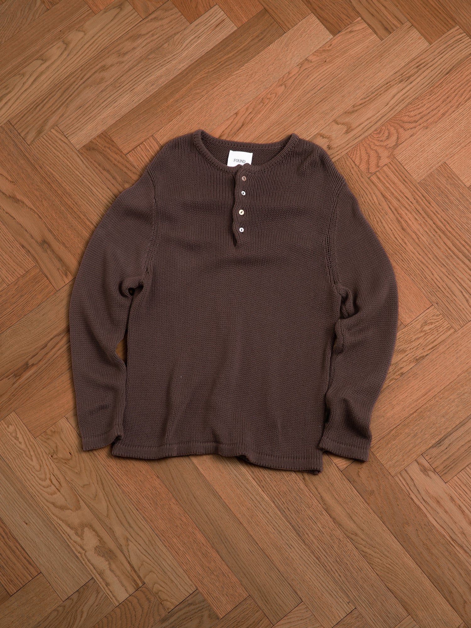 A brown Knit Henley sweater from Profound laying on a wooden floor.
