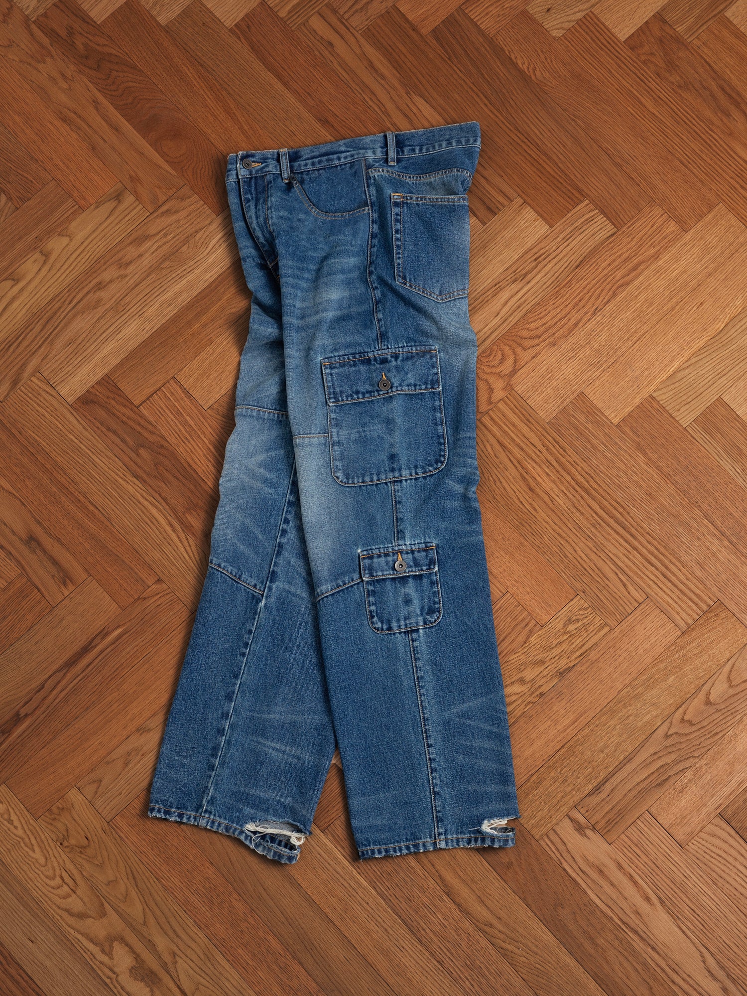 A pair of Found Siwa Cargo Paneled Jeans in blue lying flat on a herringbone patterned wooden floor.