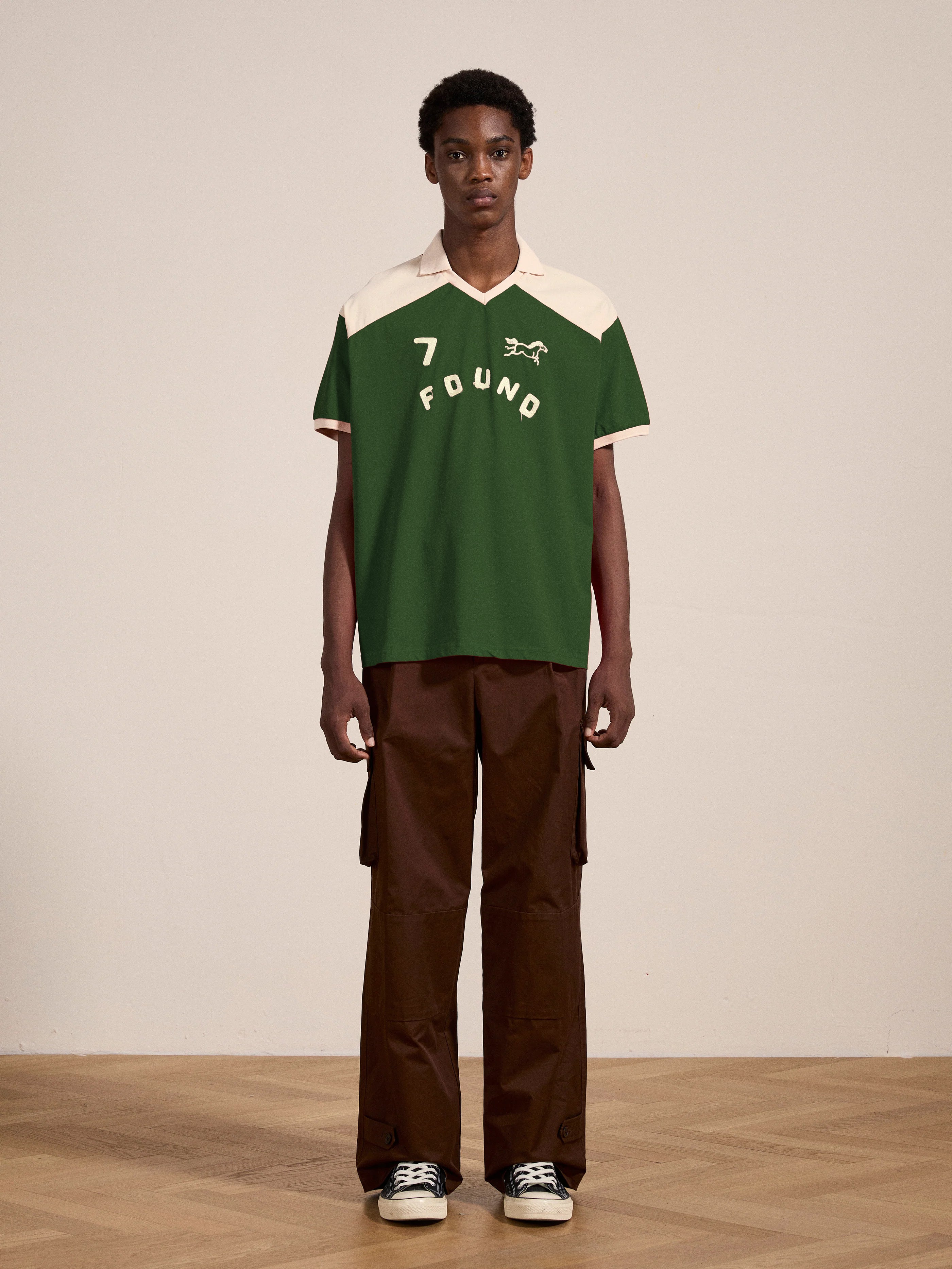 A person stands indoors on a wooden floor wearing the FOUND Goji Green Jersey, a green and white collared shirt with "7 FOUND" printed on it, paired with brown pants and black sneakers.