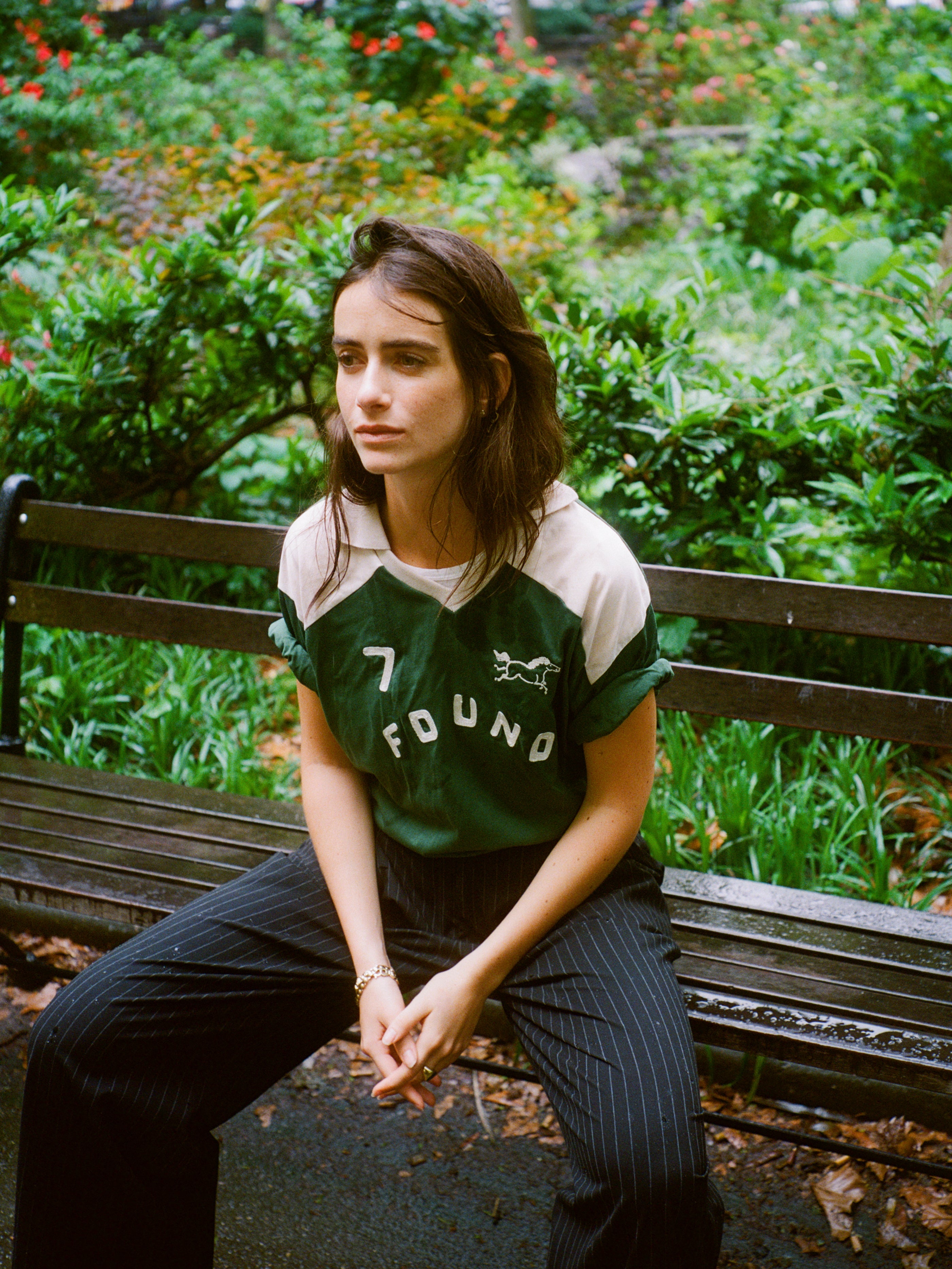 A woman with brown hair sits on a park bench, wearing the FOUND Goji Green Jersey and pinstripe pants. Green bushes and flowers fill the background.