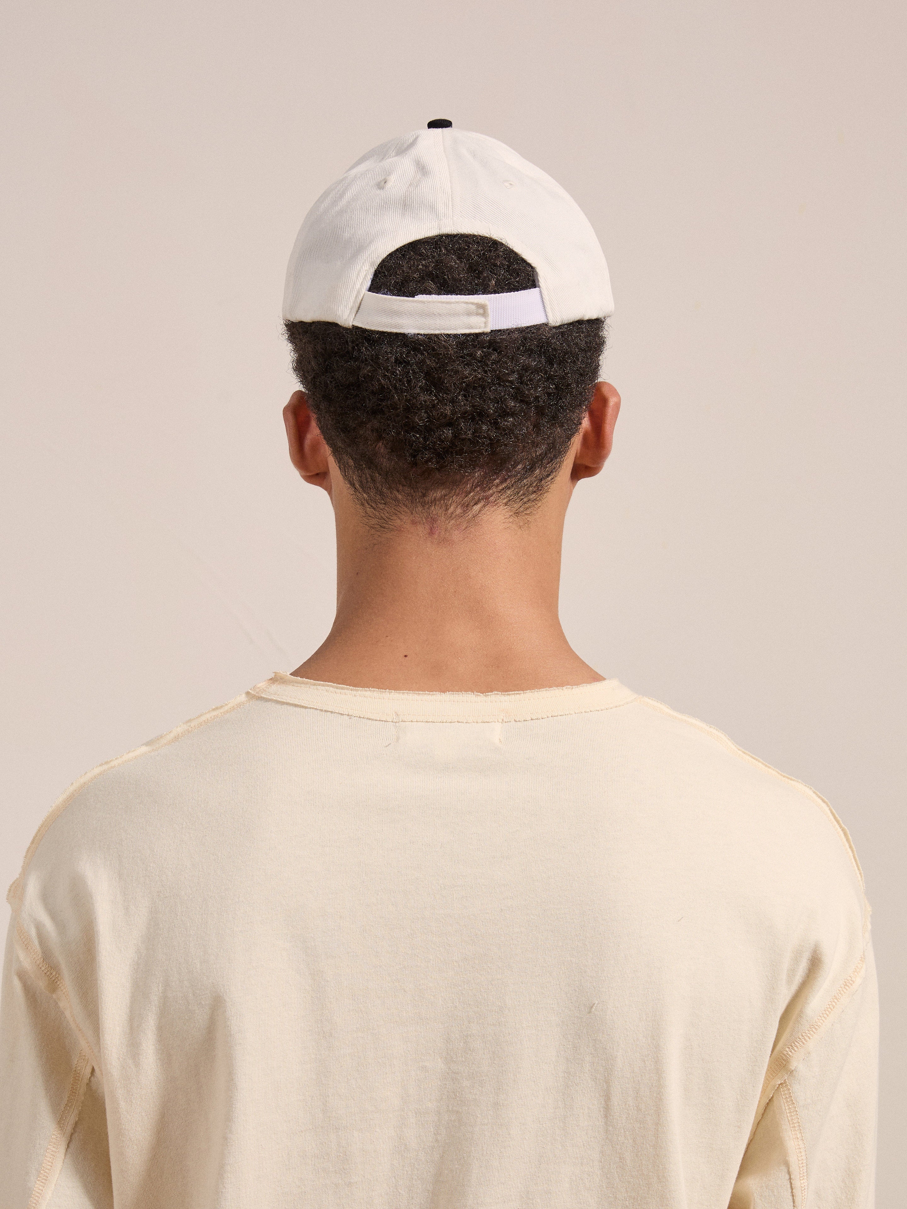 A person with short curly hair is wearing a Greenlands Contrast Cap by FOUND backward and a beige shirt. The individual is facing away from the camera.