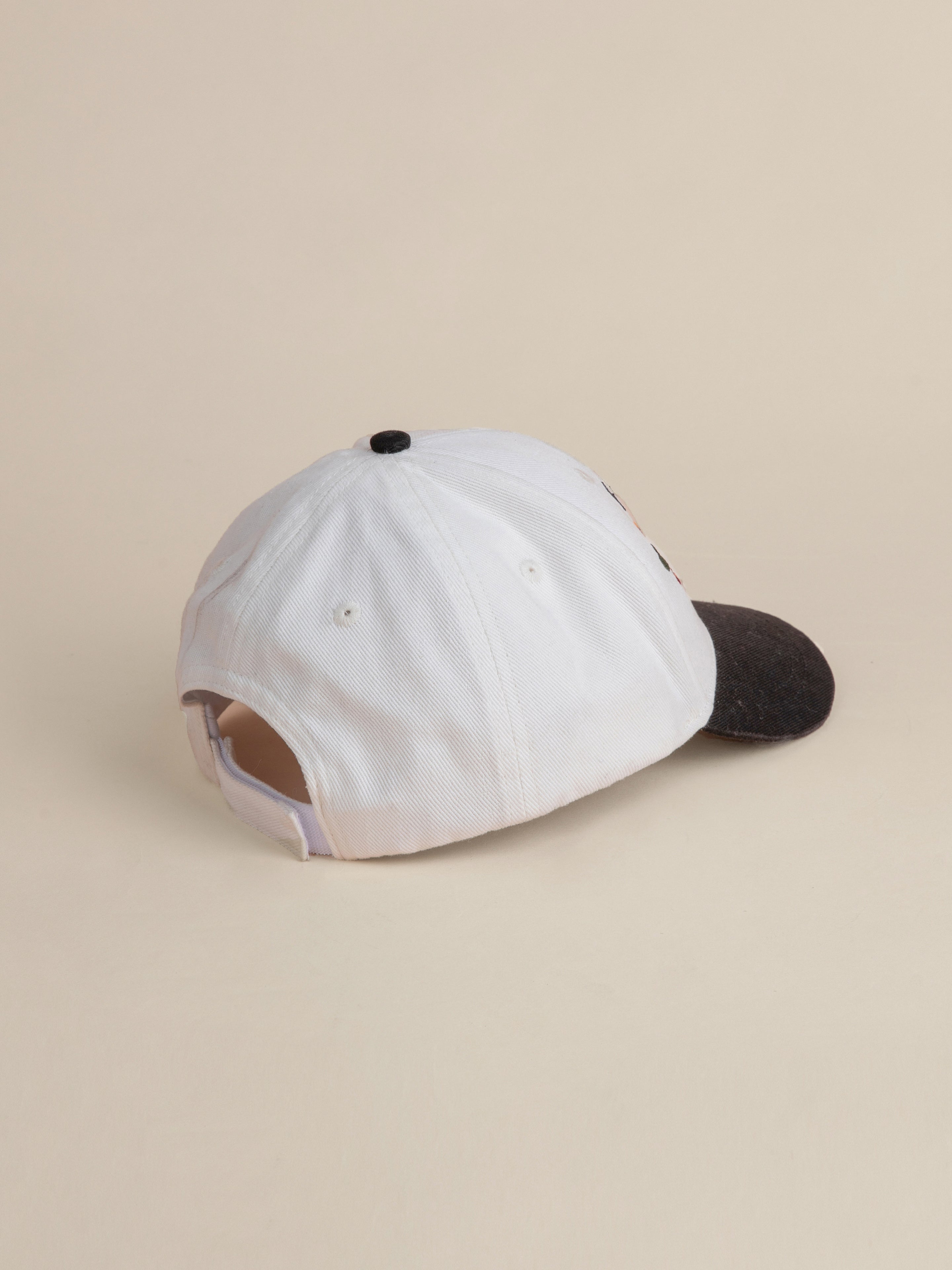 The Greenlands Contrast Cap by FOUND is displayed from the back. This white cap with a black 6-panel curved brim showcases an adjustable strap, metal eyelets for ventilation, and unique Kantha-style embroidery.