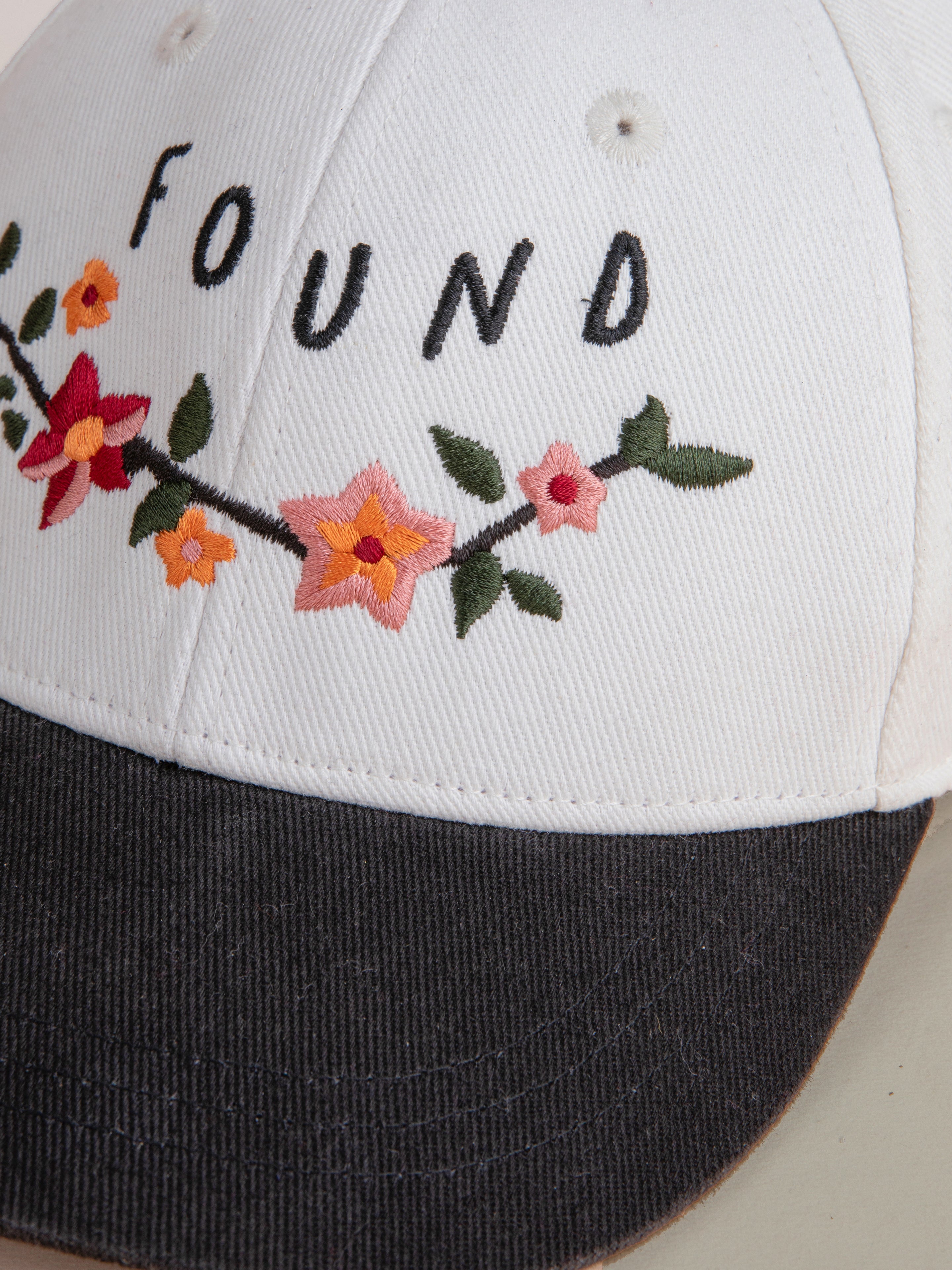 Close-up of a white cotton Greenlands Contrast Cap by FOUND, featuring Kantha-style floral embroidery on the front and a dark-colored brim.