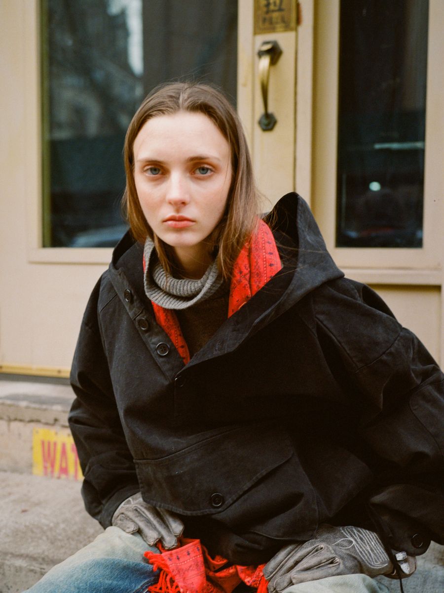 A person with long hair is seated on a step in front of a beige door, wearing a dark coat and scarf. Their timeless style is highlighted by the FOUND Washed Twill Pocket Anorak Jacket.