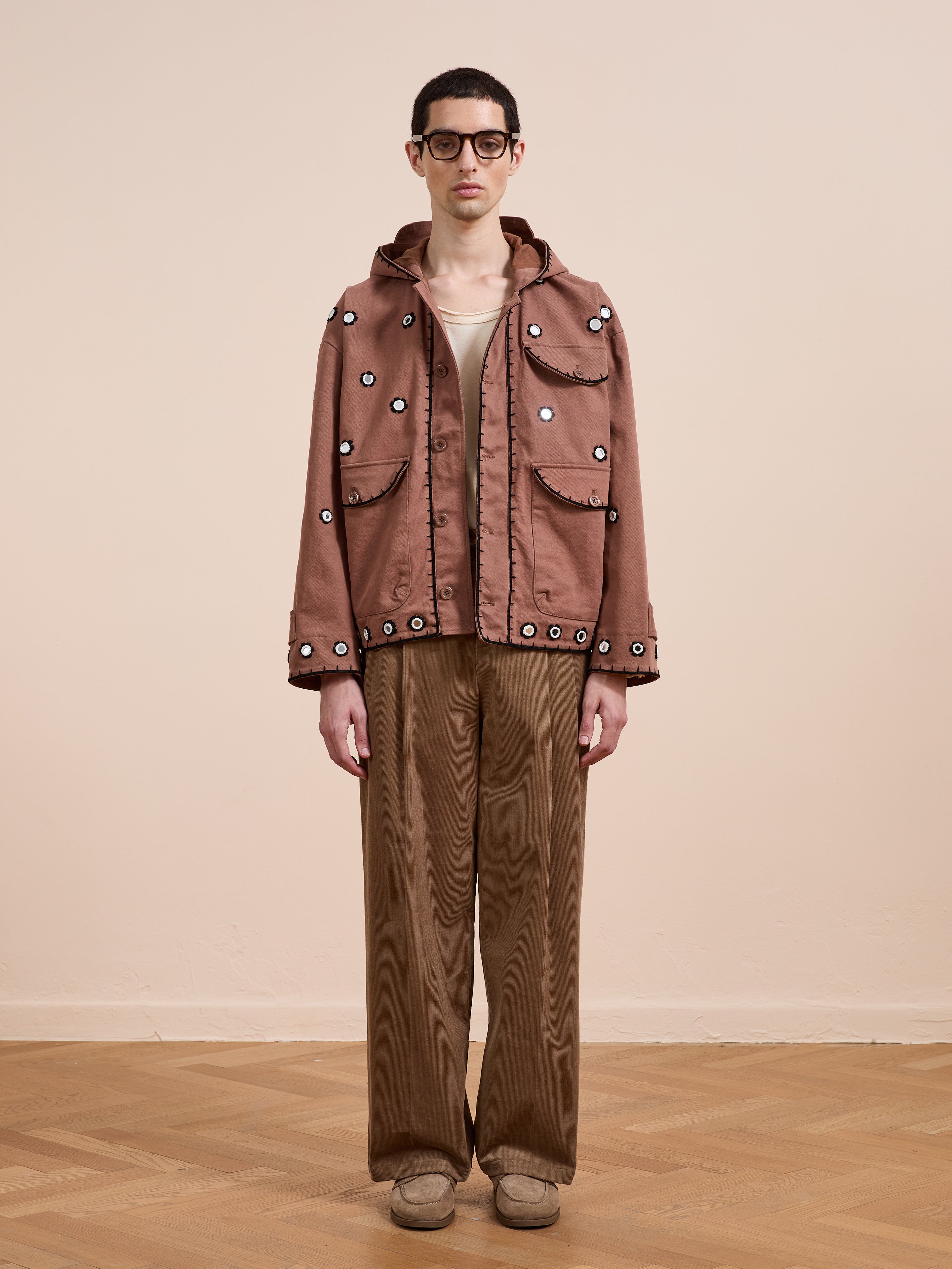 Indoors, a person wears glasses and the FOUND Hand Embroidered Hooded Mirror Anorak in brown cotton twill, paired with a beige top, brown wide-leg pants, and tan shoes on a wooden floor.