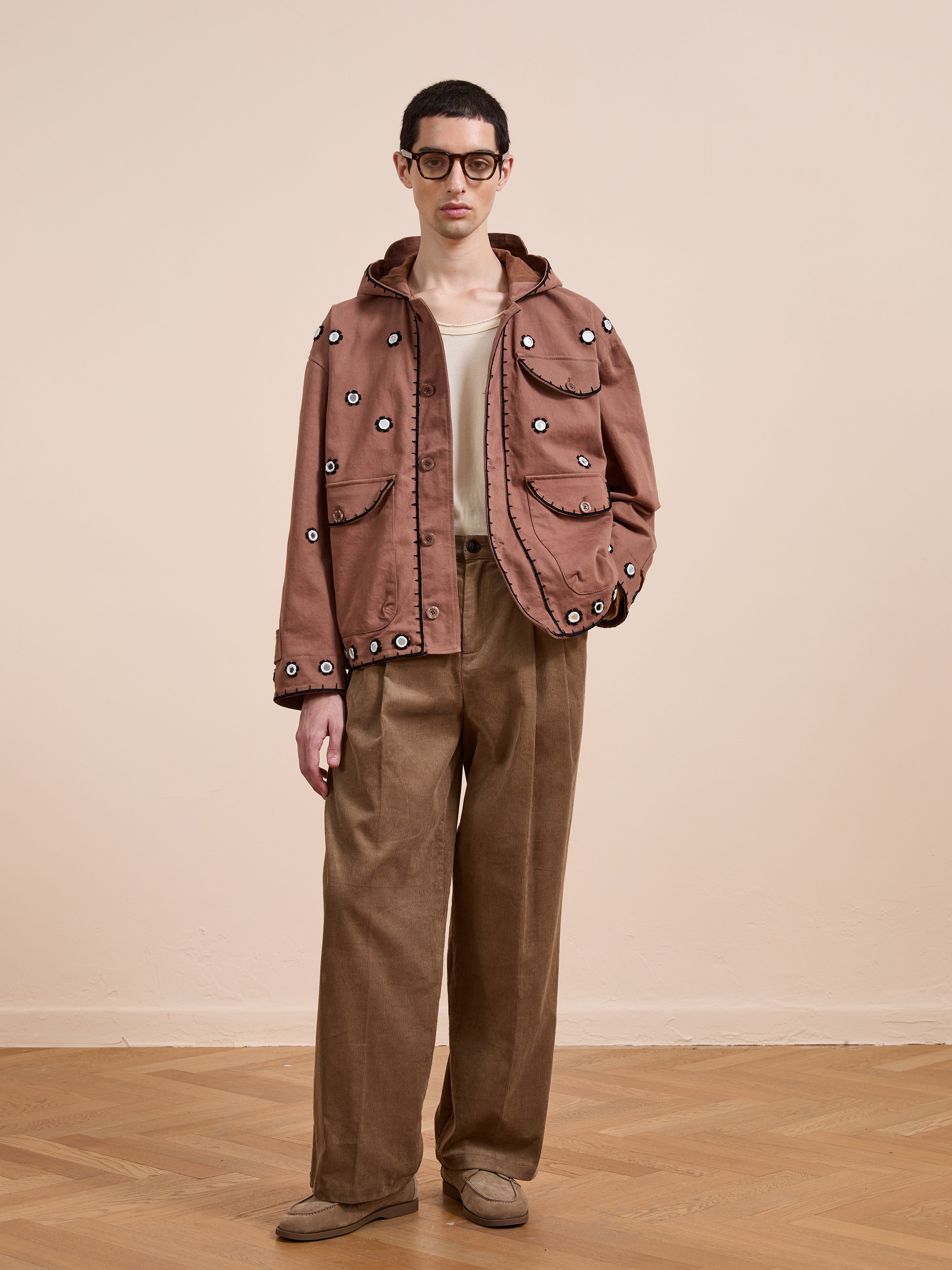 Indoors, a person wears FOUND’s Hand Embroidered Hooded Mirror Anorak with glasses, a beige shirt, wide-legged brown pants, and brown shoes.