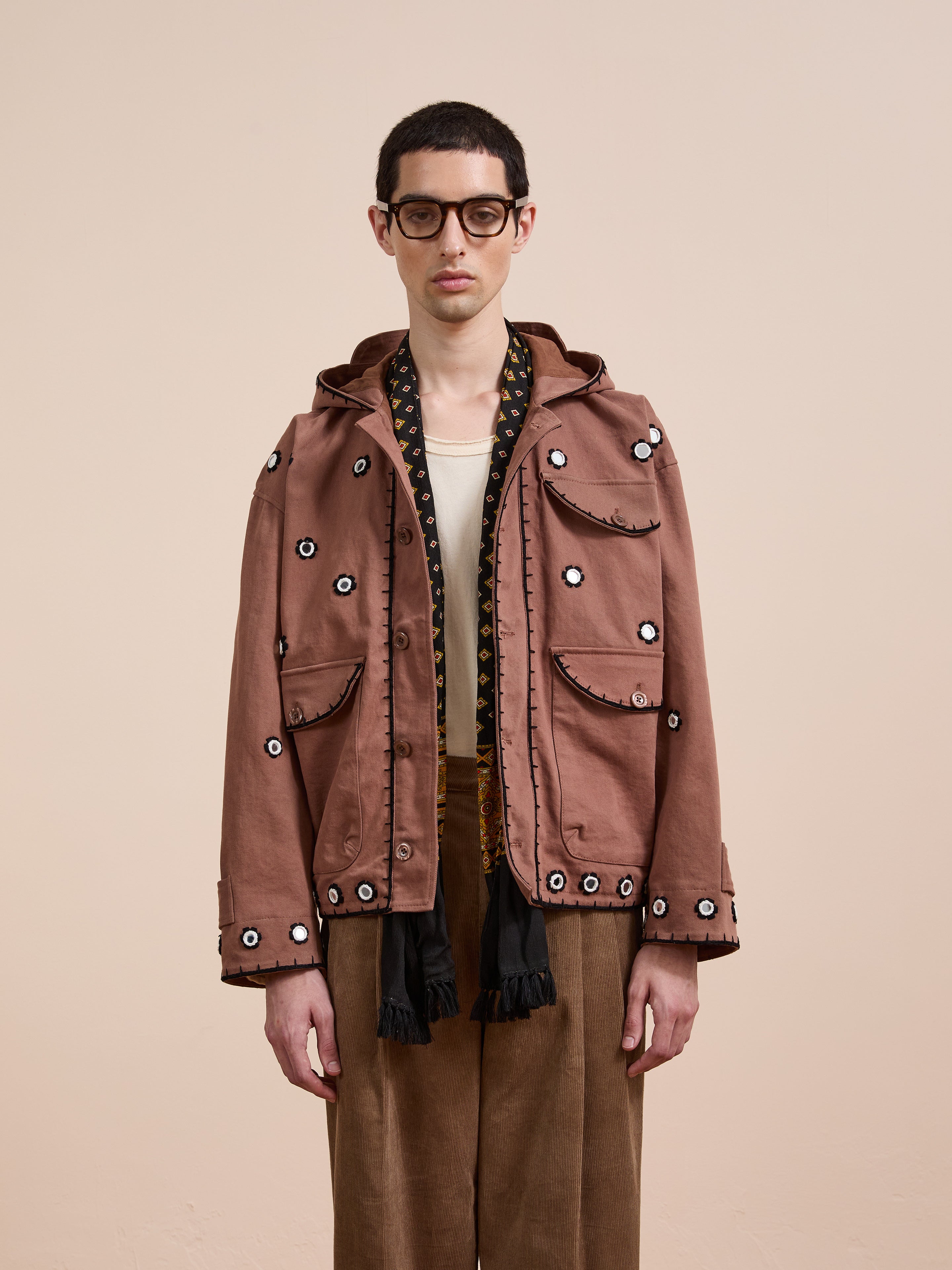 A person wearing glasses and the FOUND Hand Embroidered Hooded Mirror Anorak stands against a plain beige background, styled with a cream shirt and brown trousers.
