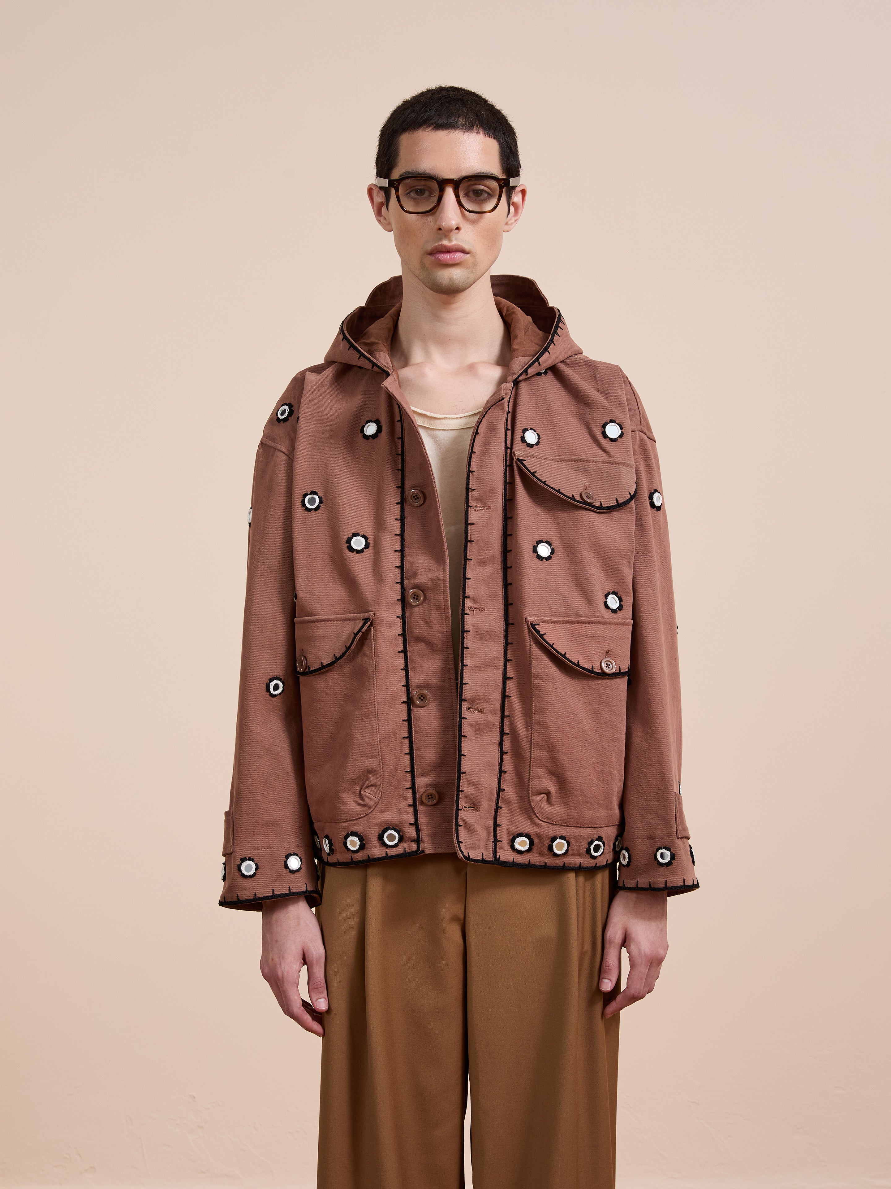 Against a plain beige background, a person wears glasses and the FOUND Hand Embroidered Hooded Mirror Anorak with matching brown pants.