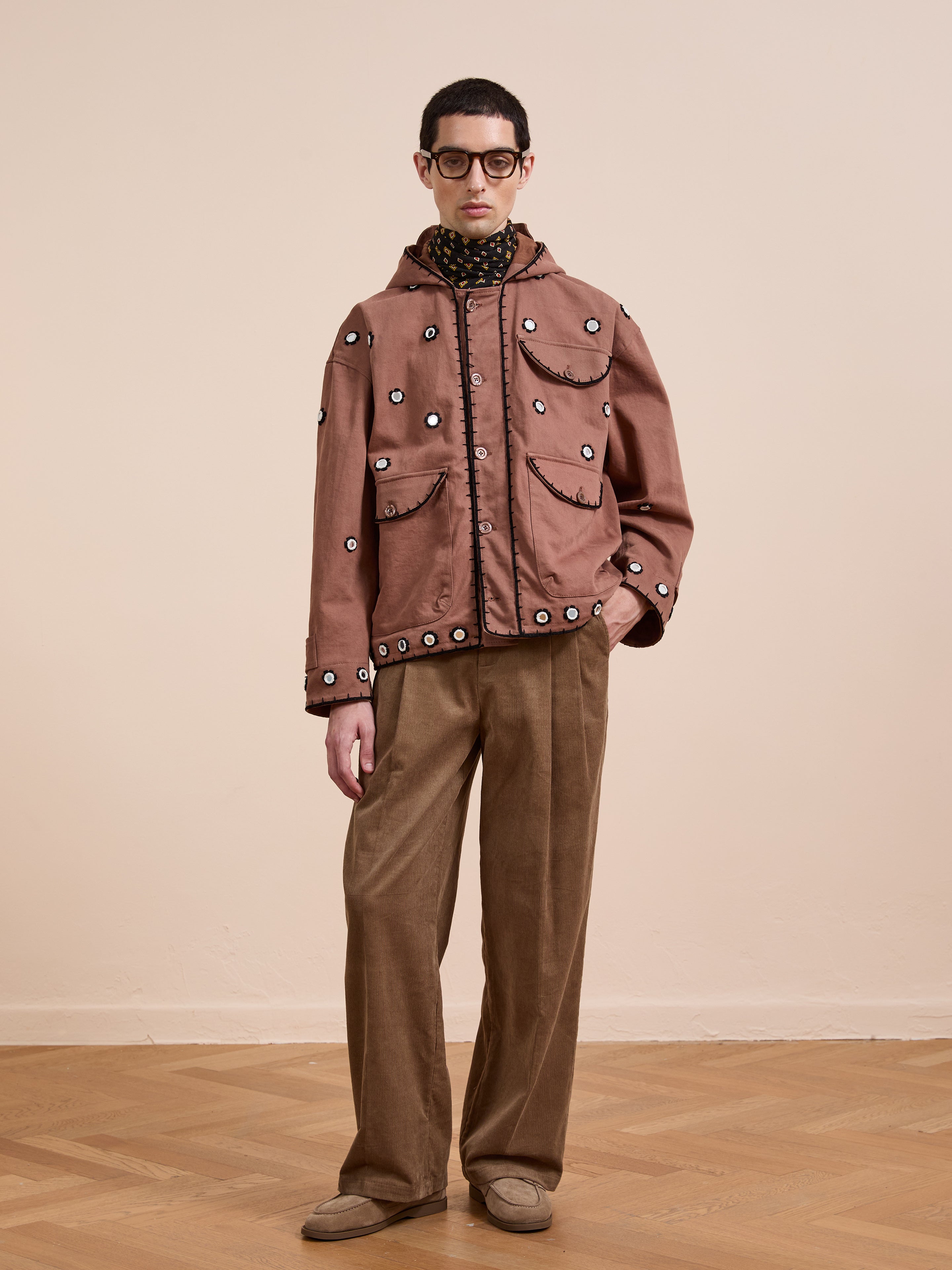 A person stands indoors against a light wall and wood floor, wearing FOUND's Hand Embroidered Hooded Mirror Anorak, glasses, a patterned scarf, tan wide-leg pants, and beige shoes.