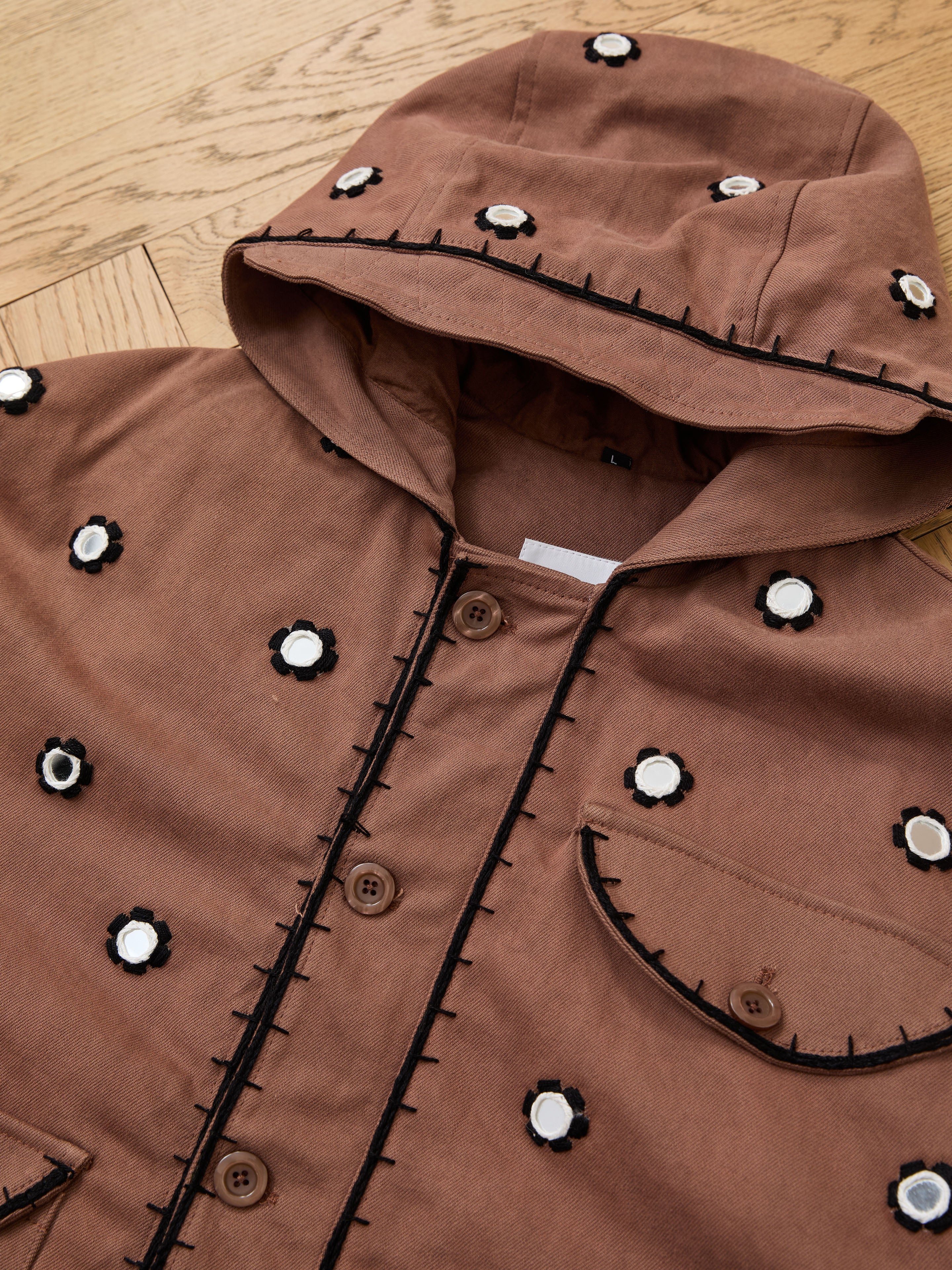 The FOUND Hand Embroidered Hooded Mirror Anorak features brown fabric with white and black circular mirror embroidery and black stitching, laid flat on wood to highlight exquisite South Asian craftsmanship.