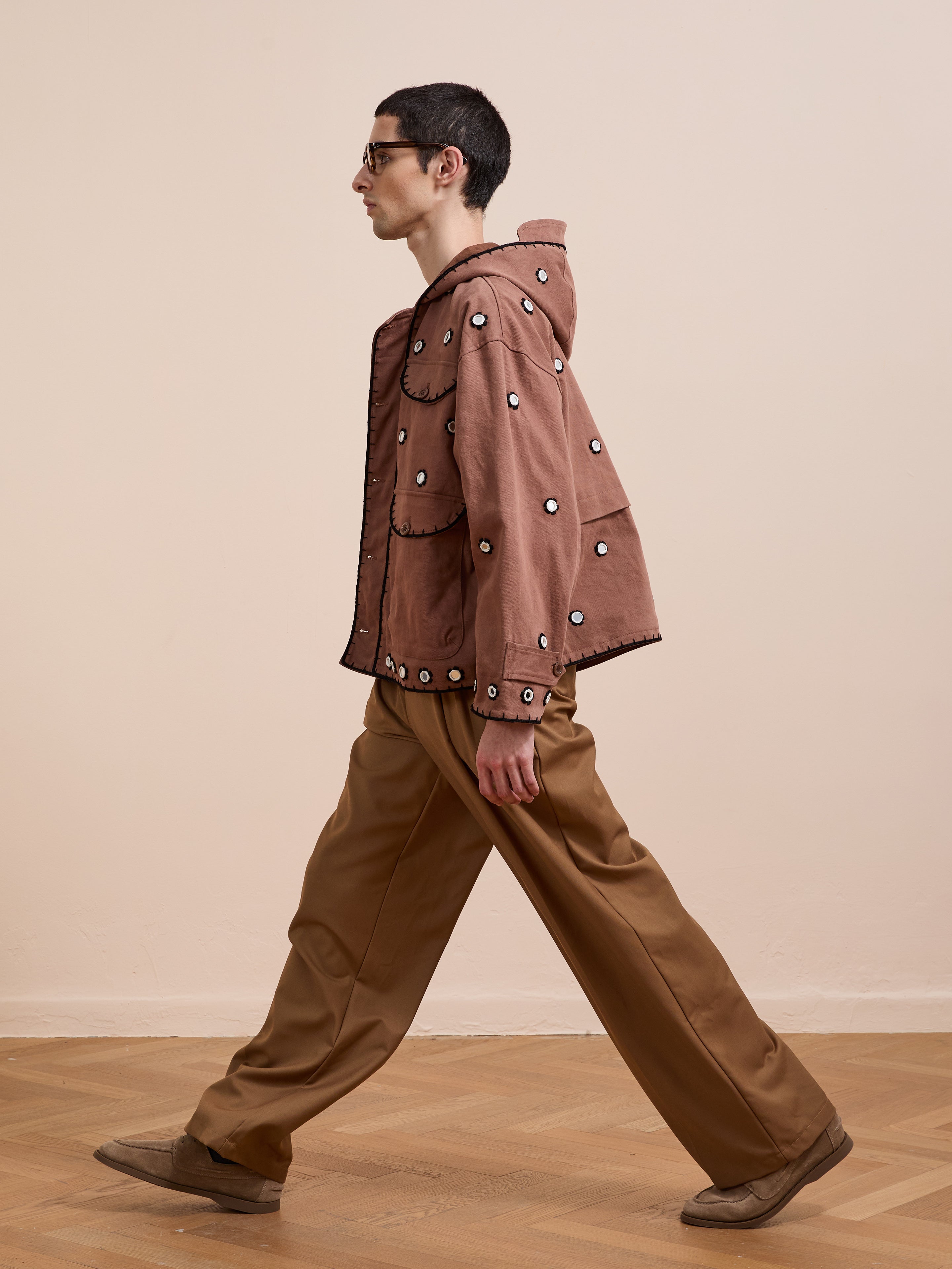A person with short dark hair and glasses wears the FOUND Hand Embroidered Hooded Mirror Anorak over tan wide-leg pants and beige shoes, standing indoors against a plain, light-colored background.