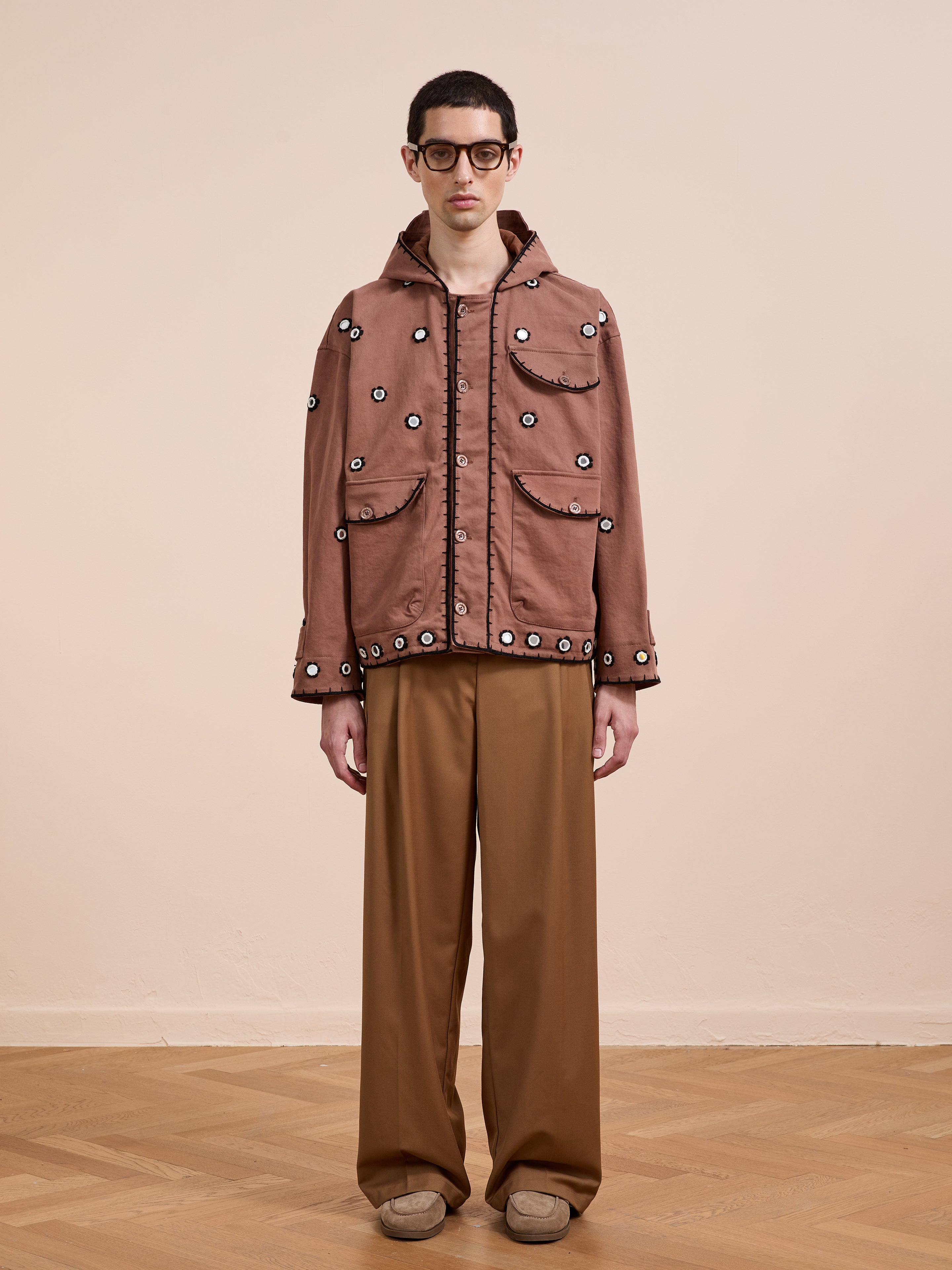 A person in FOUND's Hand Embroidered Hooded Mirror Anorak with multiple pockets and brown wide-leg trousers stands on a wooden floor against a beige wall, wearing black glasses.