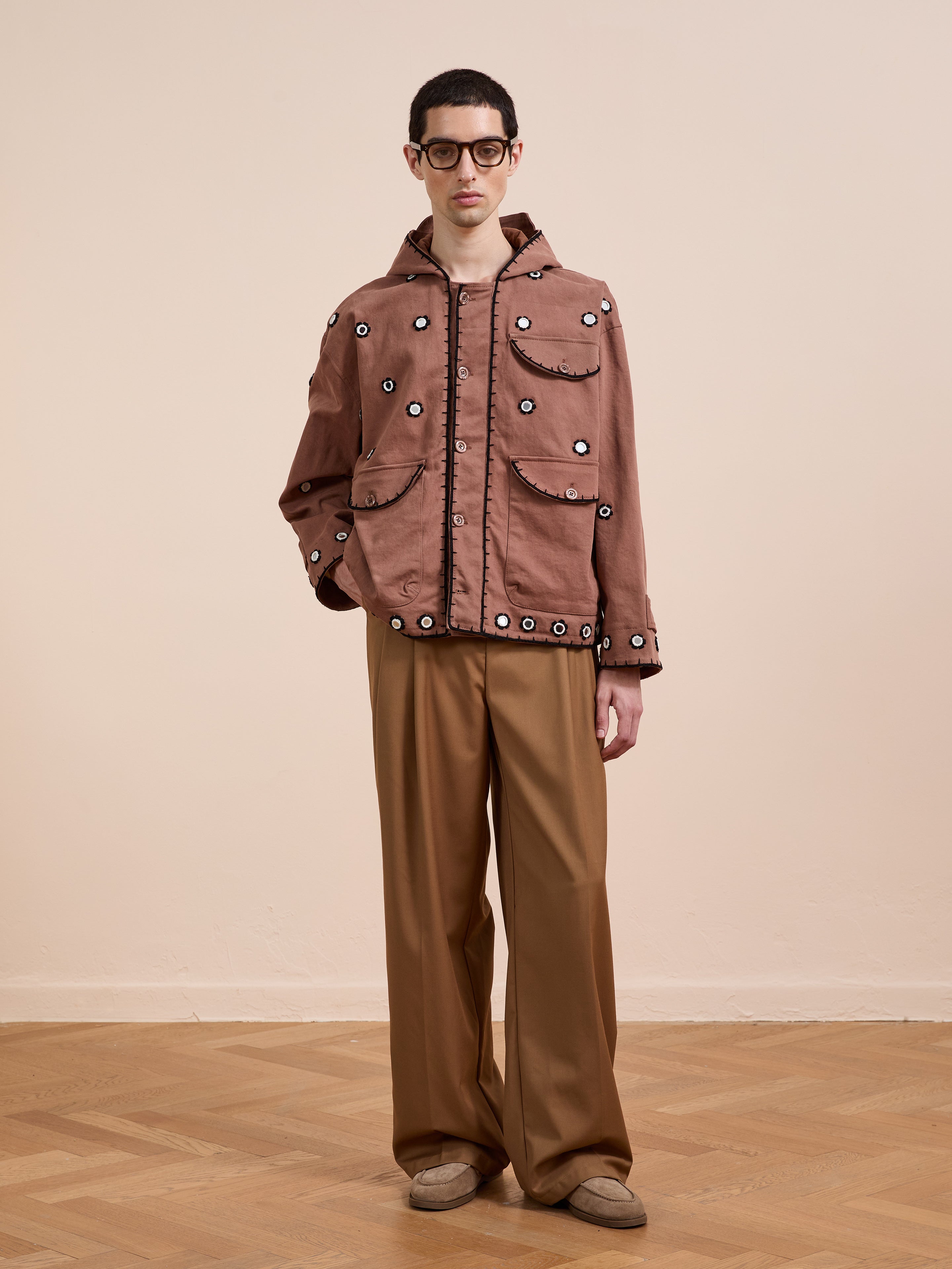 Indoors, a person wears the FOUND Hand Embroidered Hooded Mirror Anorak with front pockets, paired with loose tan trousers, glasses, and beige shoes on a wooden floor.