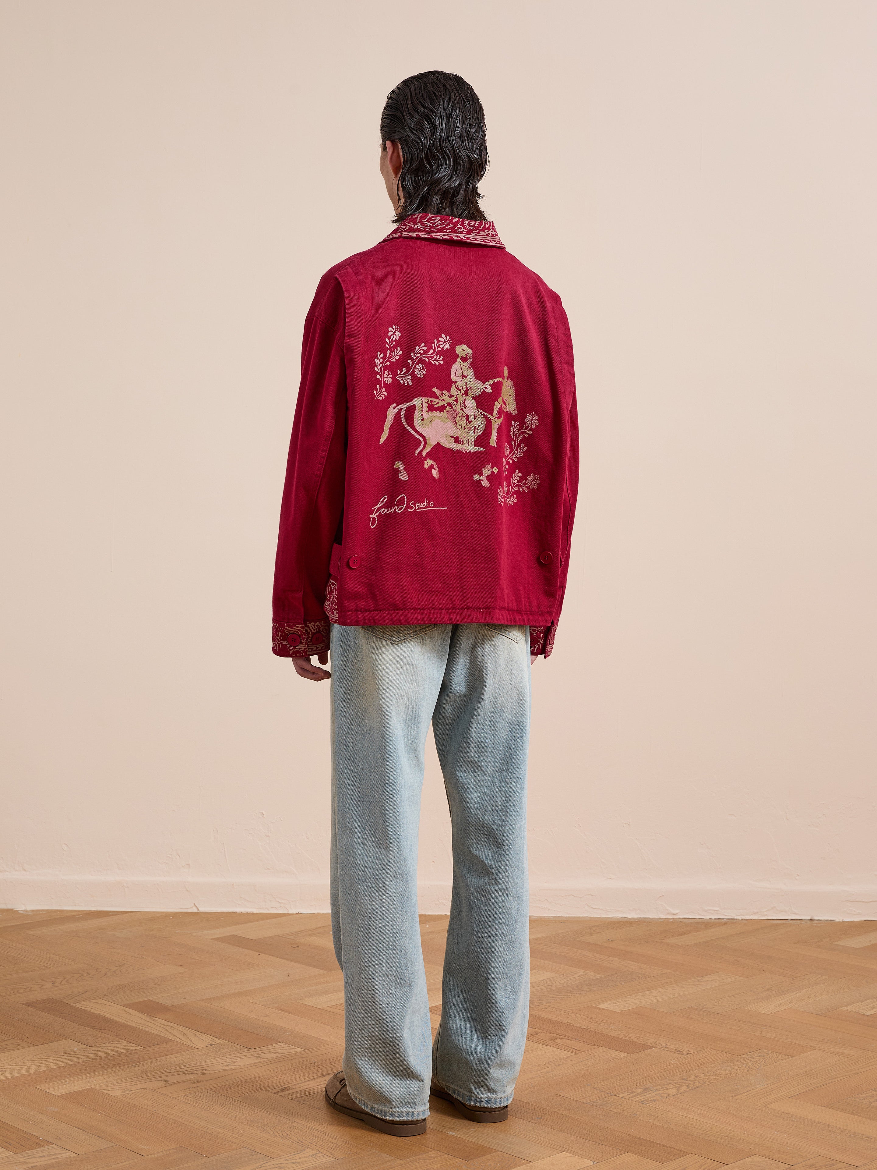 Someone stands facing away in a Found Abhala Printed Work Jacket with equestrian motifs and floral embroidery on the back, paired with light blue jeans and brown shoes on a wooden floor.
