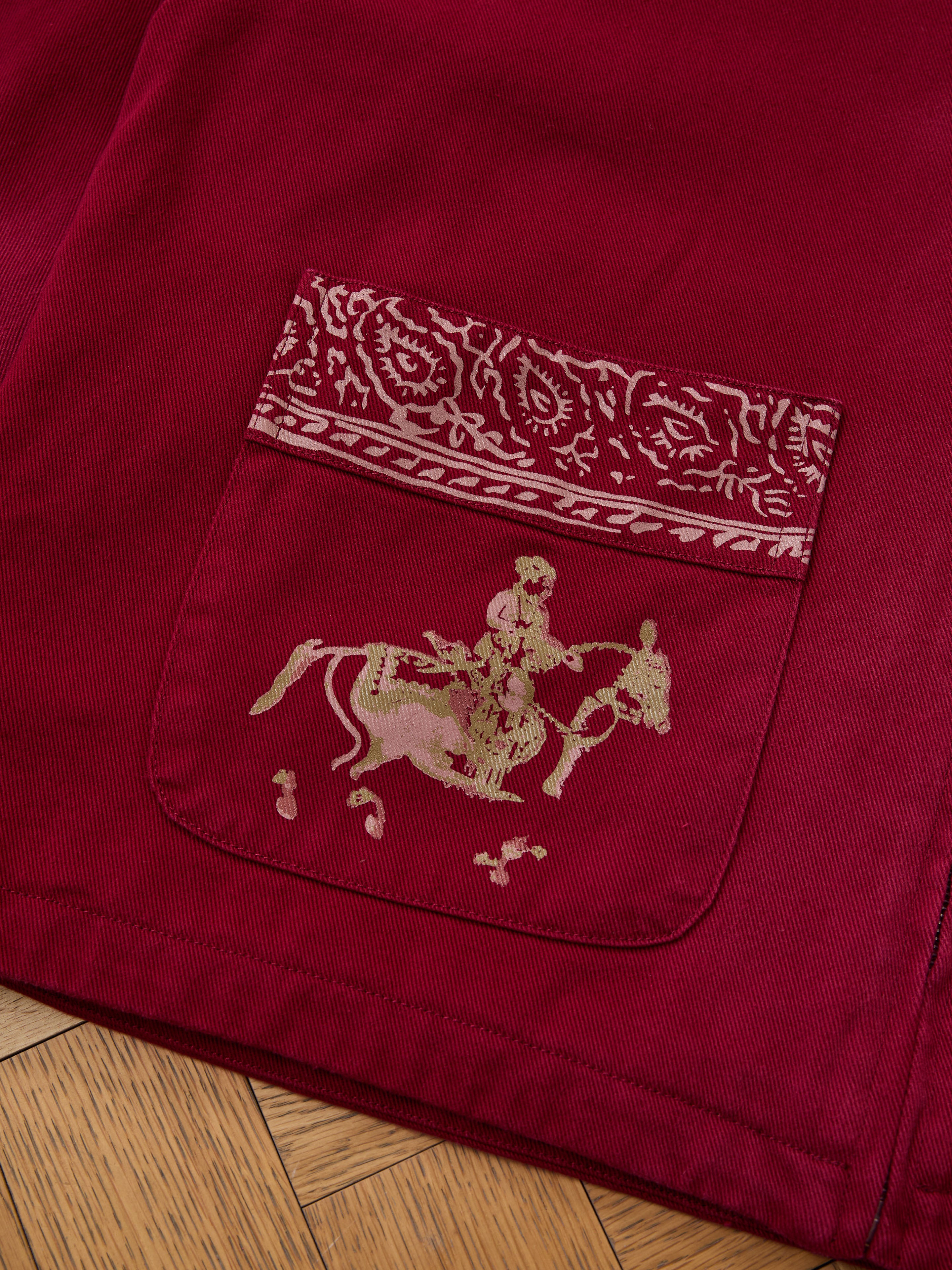 Close-up of Found's Abhala Printed Work Jacket pocket—red, unisex, with a gold horse rider and equestrian motifs. A patterned strip accents the top edge.