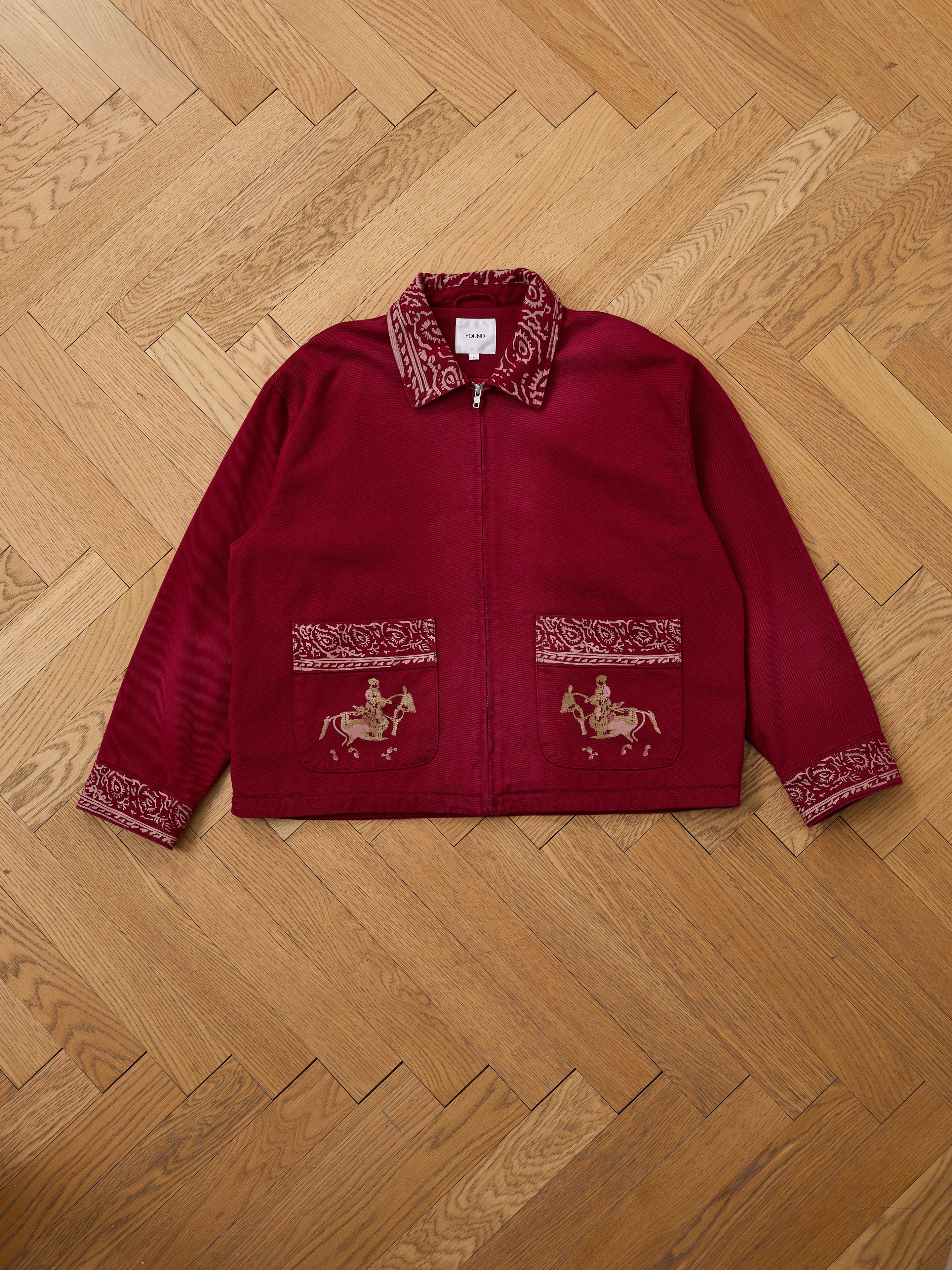 A Found Abhala Printed Work Jacket in burgundy, featuring a zip-up front, patterned collar and pockets with equestrian motifs, is laid flat on a wooden herringbone floor.