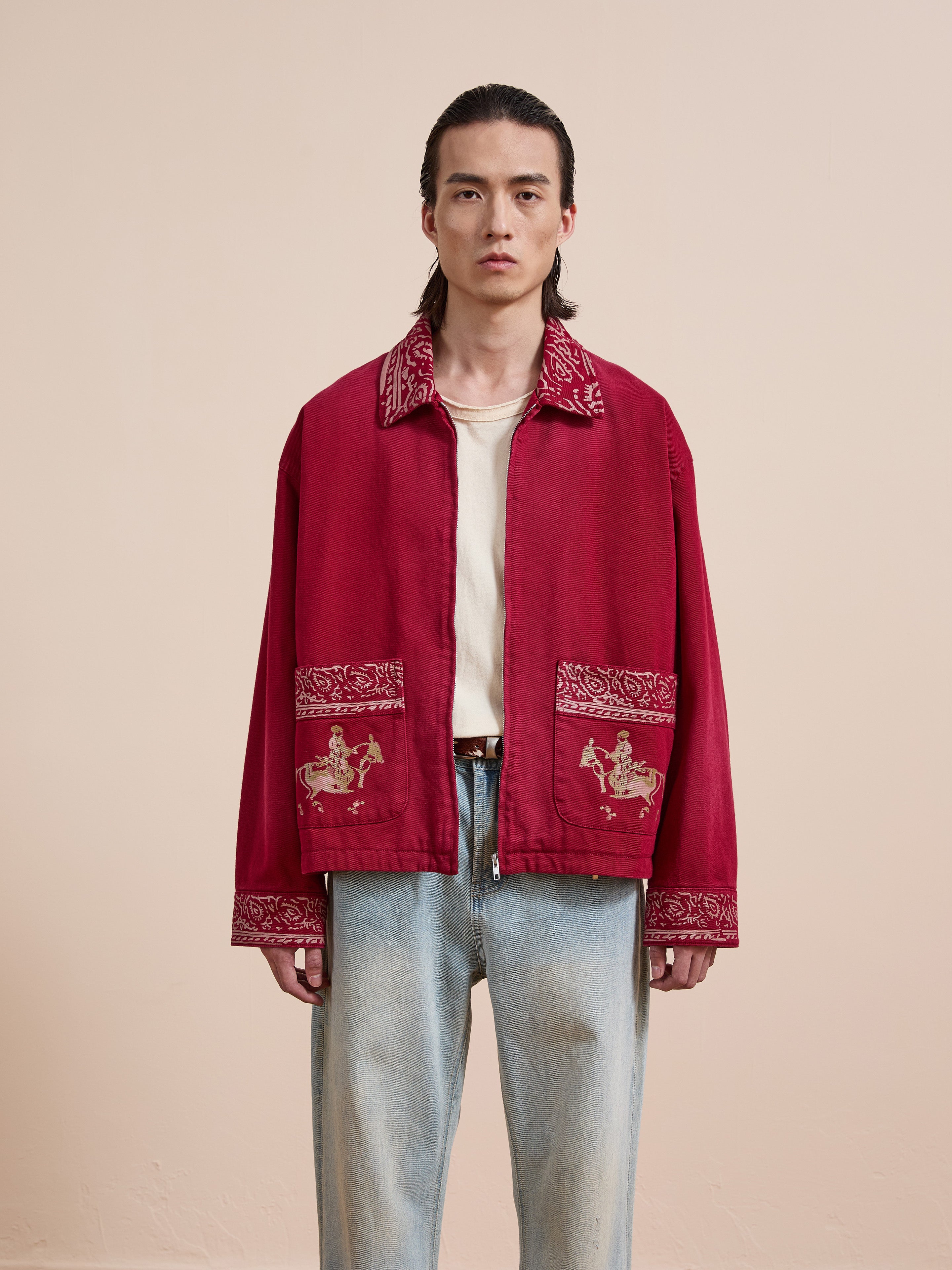 A person stands facing forward in the Found Abhala Printed Work Jacket with equestrian motifs and light blue jeans, set against a plain beige background; this unisex relaxed fit style adds a unique touch to the look.