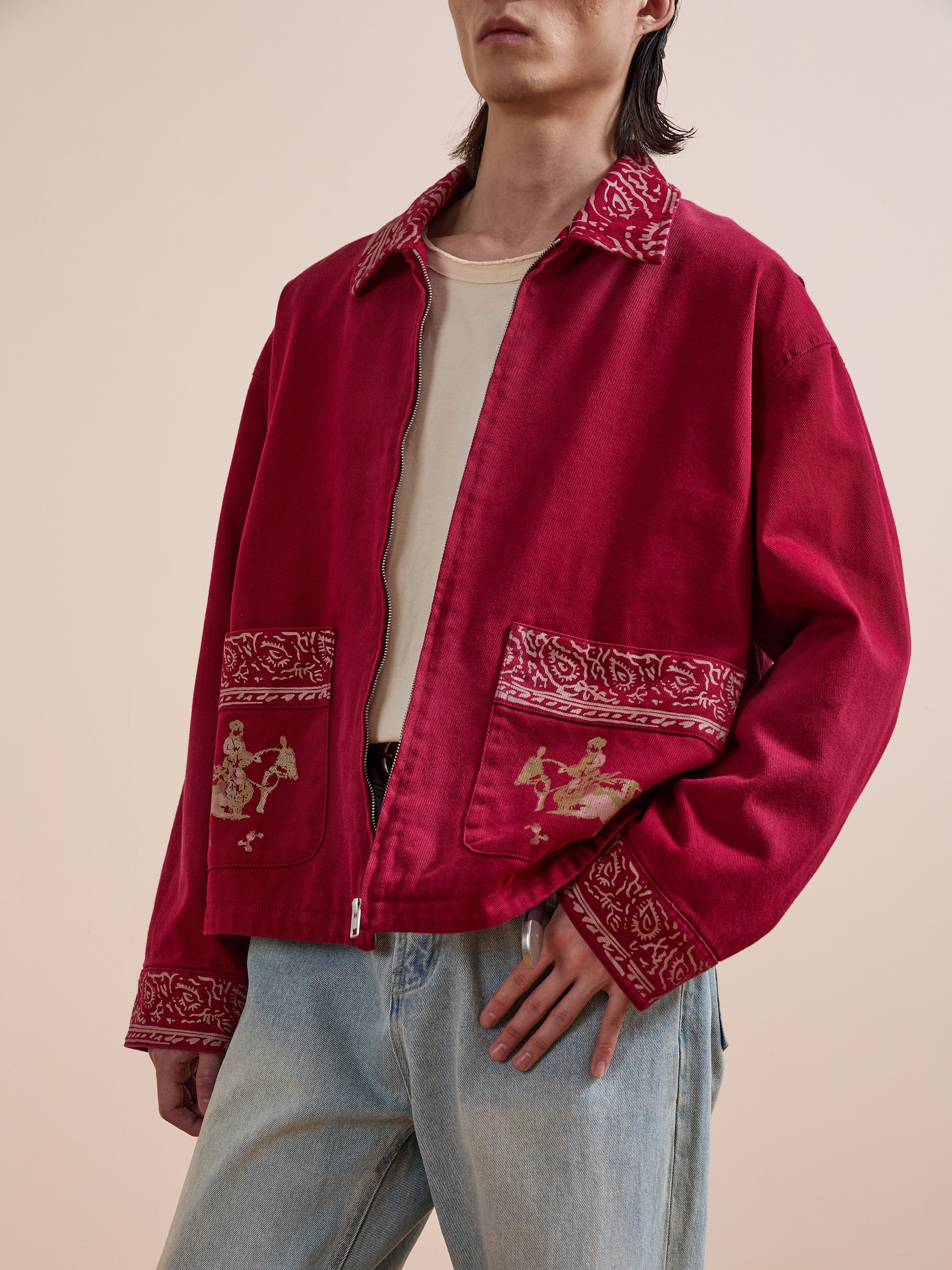 A person wears the Found Abhala Printed Work Jacket, a red unisex jacket with patterned trim and embroidered equestrian-inspired pocket designs, paired with a white shirt and light blue jeans.