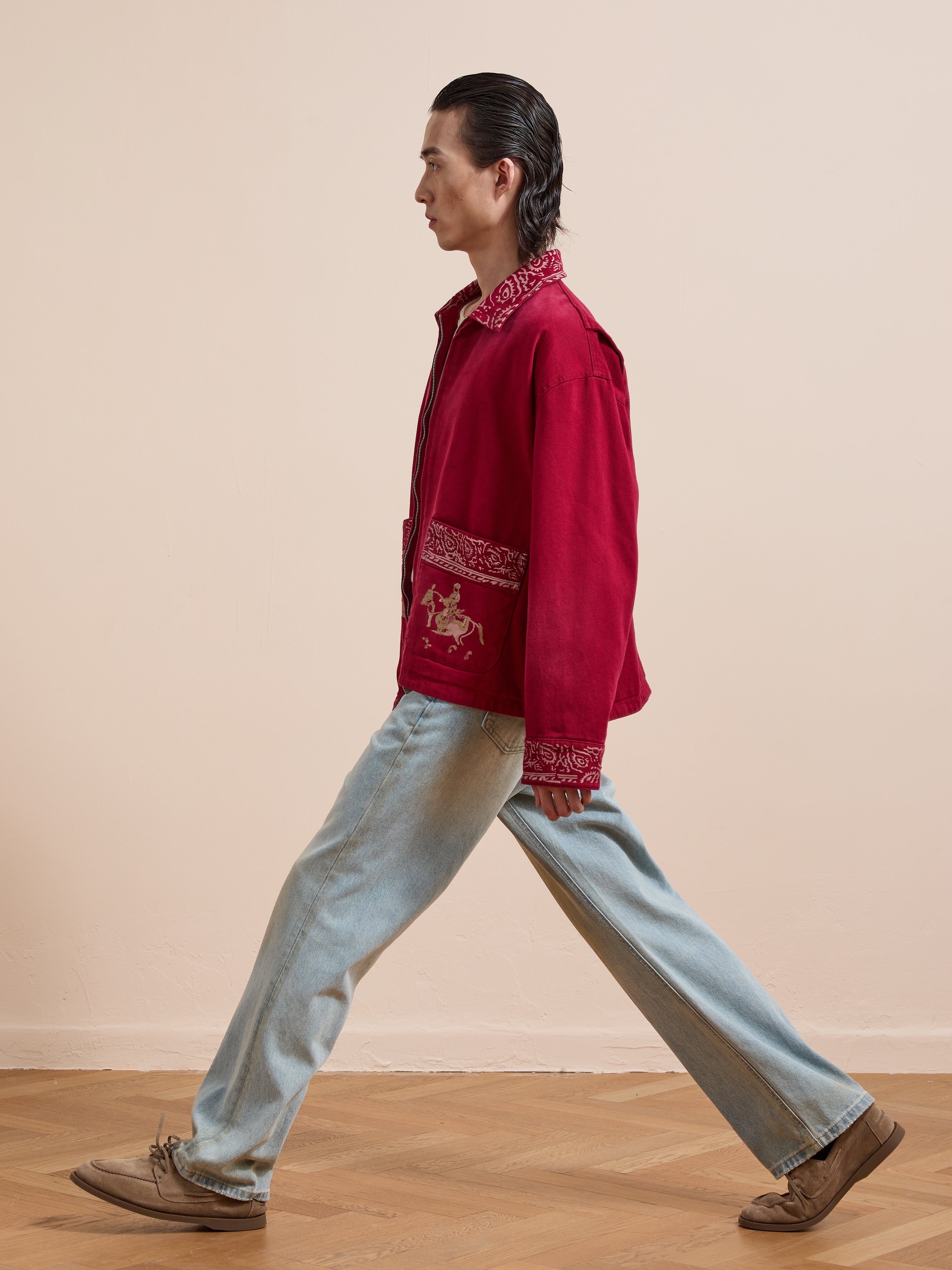 A person with slicked-back hair walks sideways, wearing the Found Abhala Printed Work Jacket with equestrian motifs, paired with light blue unisex relaxed-fit jeans and tan shoes on a wooden floor against a beige wall.