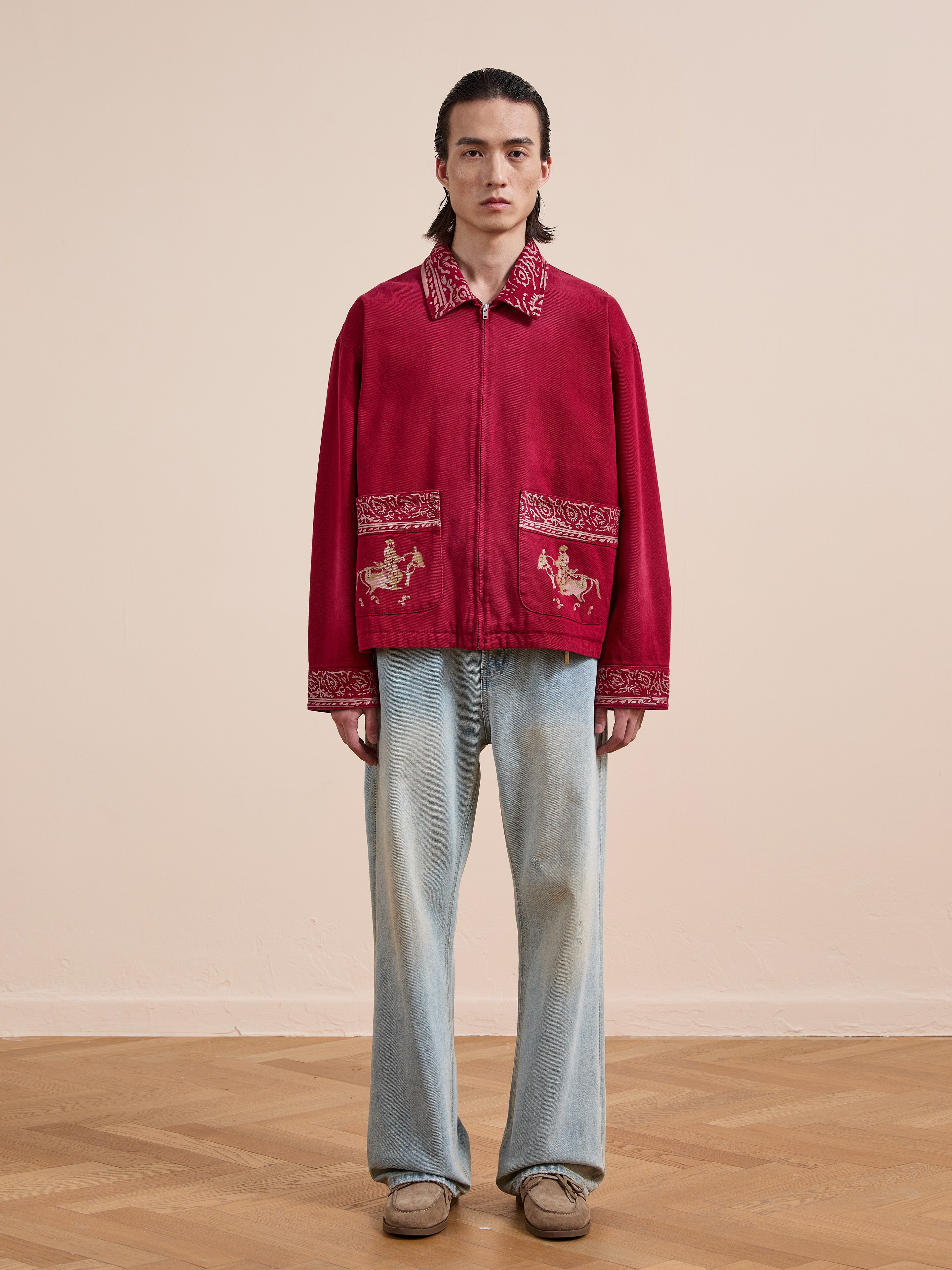 Indoors, a person wears the Found Abhala Printed Work Jacket with equestrian prints, paired with light blue jeans and beige shoes on a wooden floor. The unisex jacket offers a relaxed fit for easy, effortless style.