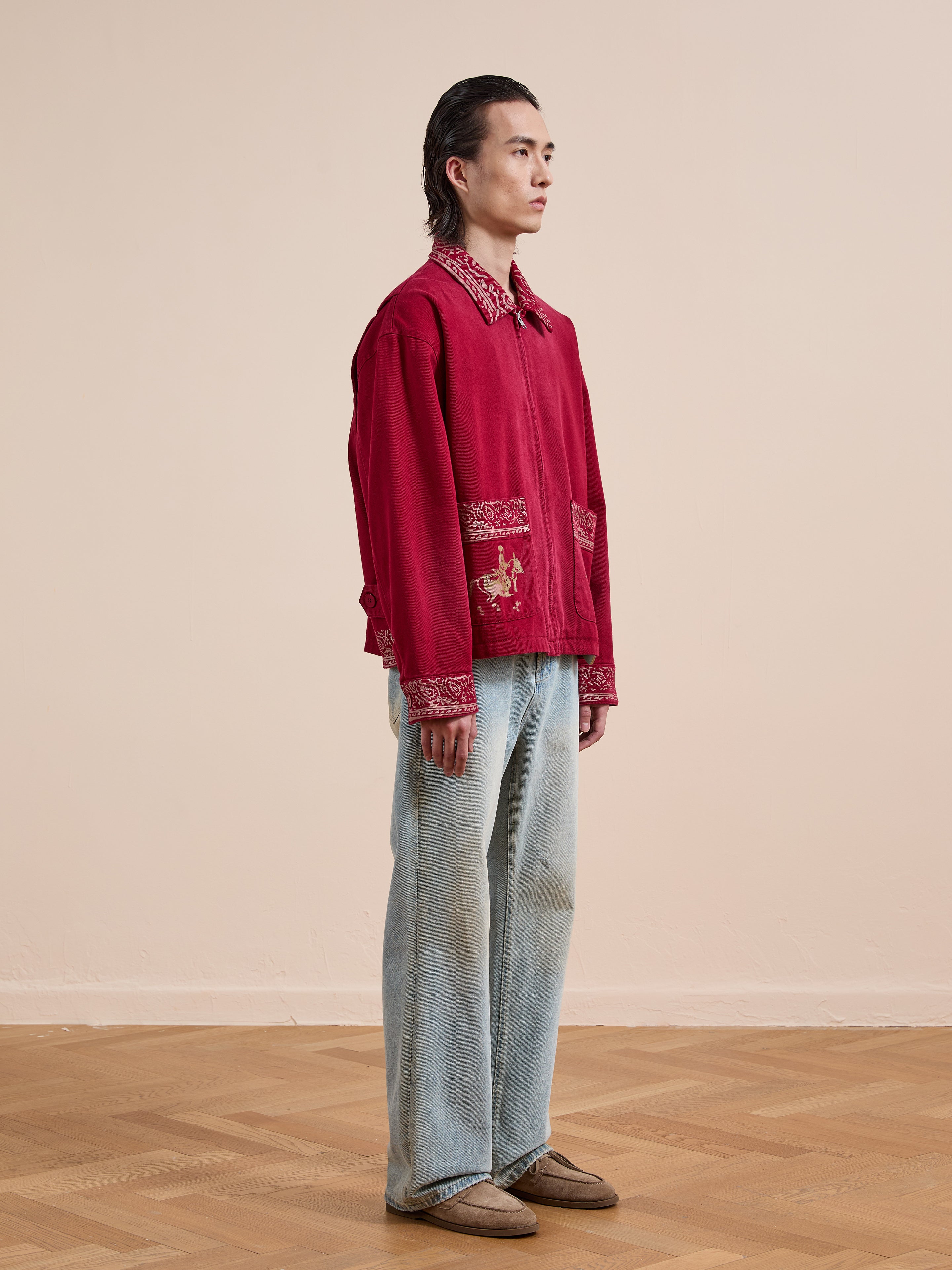 Indoors on a wooden floor against a beige wall, a person wears the Found Abhala Printed Work Jacket with equestrian motifs, paired with light blue jeans and brown shoes.