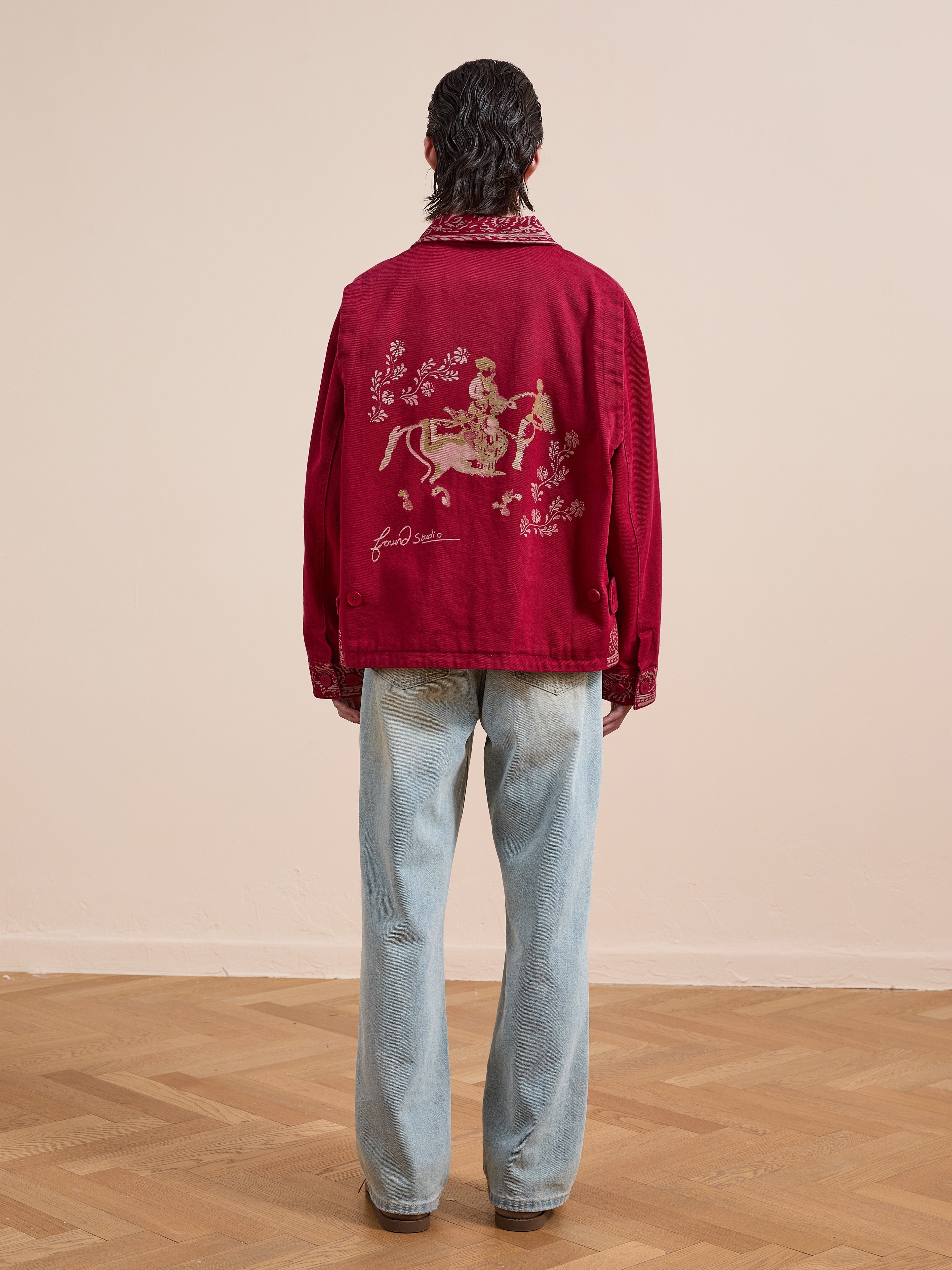 A person faces away indoors, wearing light blue jeans and the Found Abhala Printed Work Jacket—a unisex jacket featuring equestrian motifs with an embroidered person on horseback surrounded by flowers on the back.