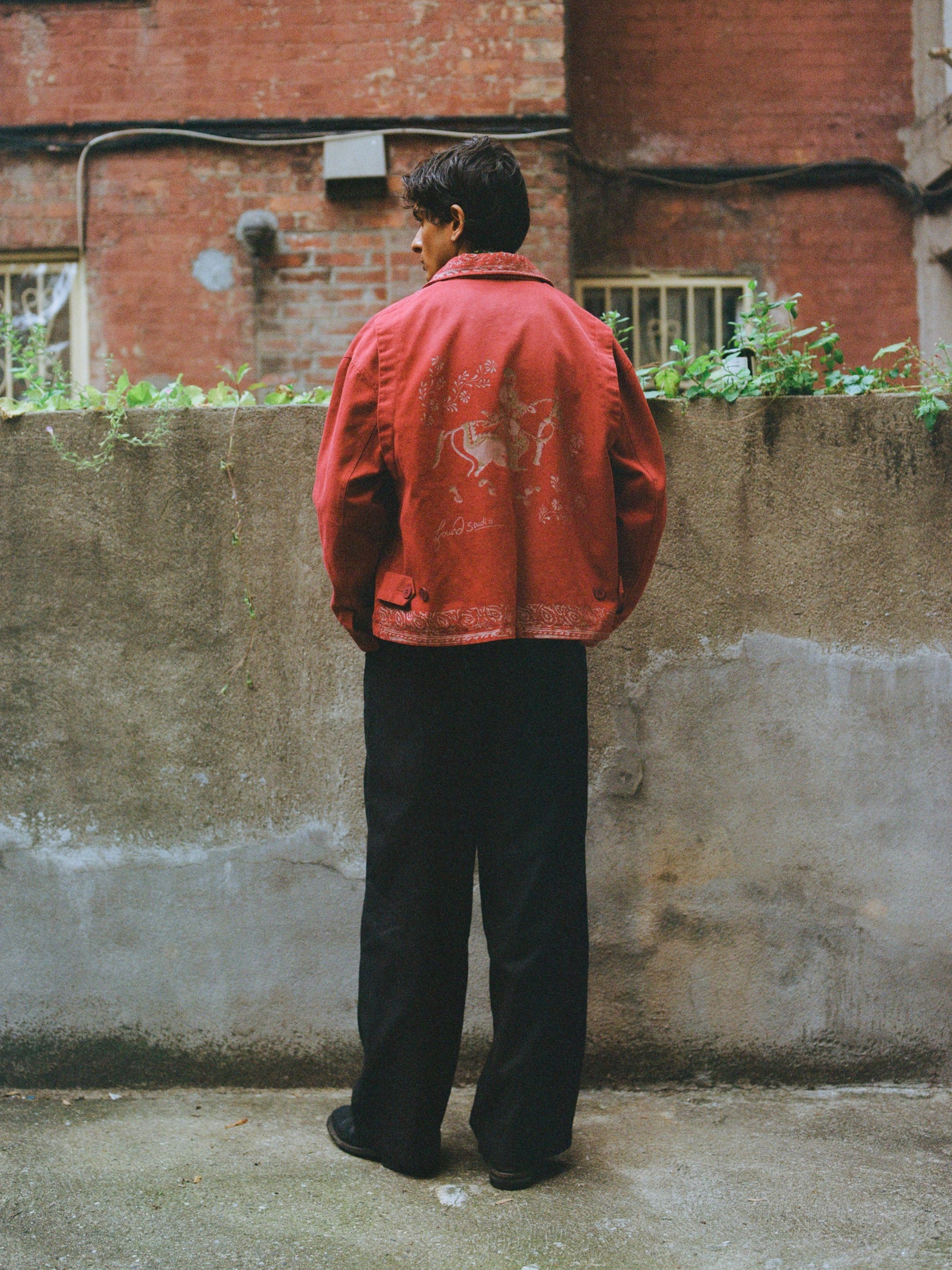 A person stands outdoors facing a concrete wall, wearing the Found Abhala Printed Work Jacket with equestrian motifs in a unisex relaxed fit and black pants, with brick buildings in the background.
