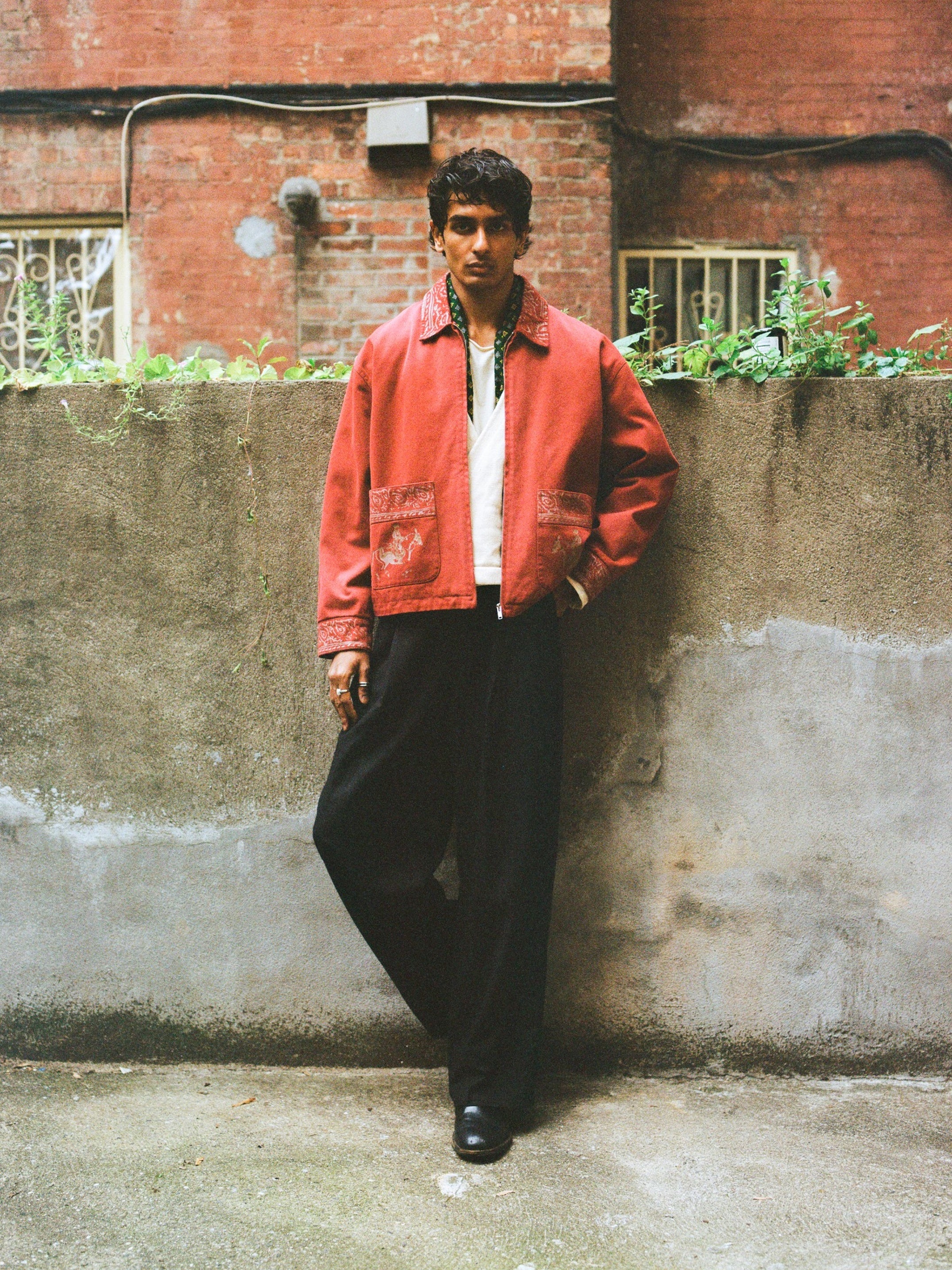 A man stands against a concrete wall in the Found Abhala Printed Work Jacket (unisex relaxed fit), paired with a white shirt and black pants, with an urban brick building behind him.