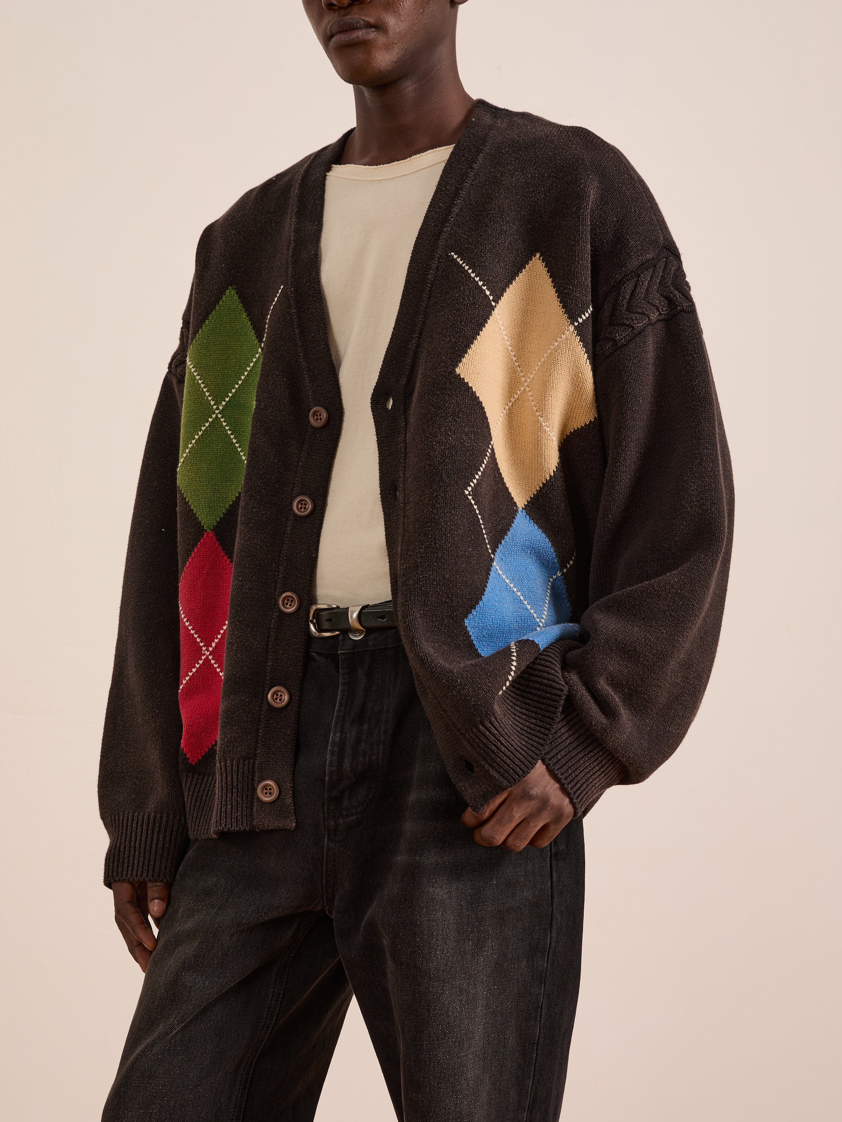 A person wears the FOUND Argyle Knit Cardigan, featuring colorful diamond patterns, paired with a cream shirt and dark jeans, standing against a neutral background.