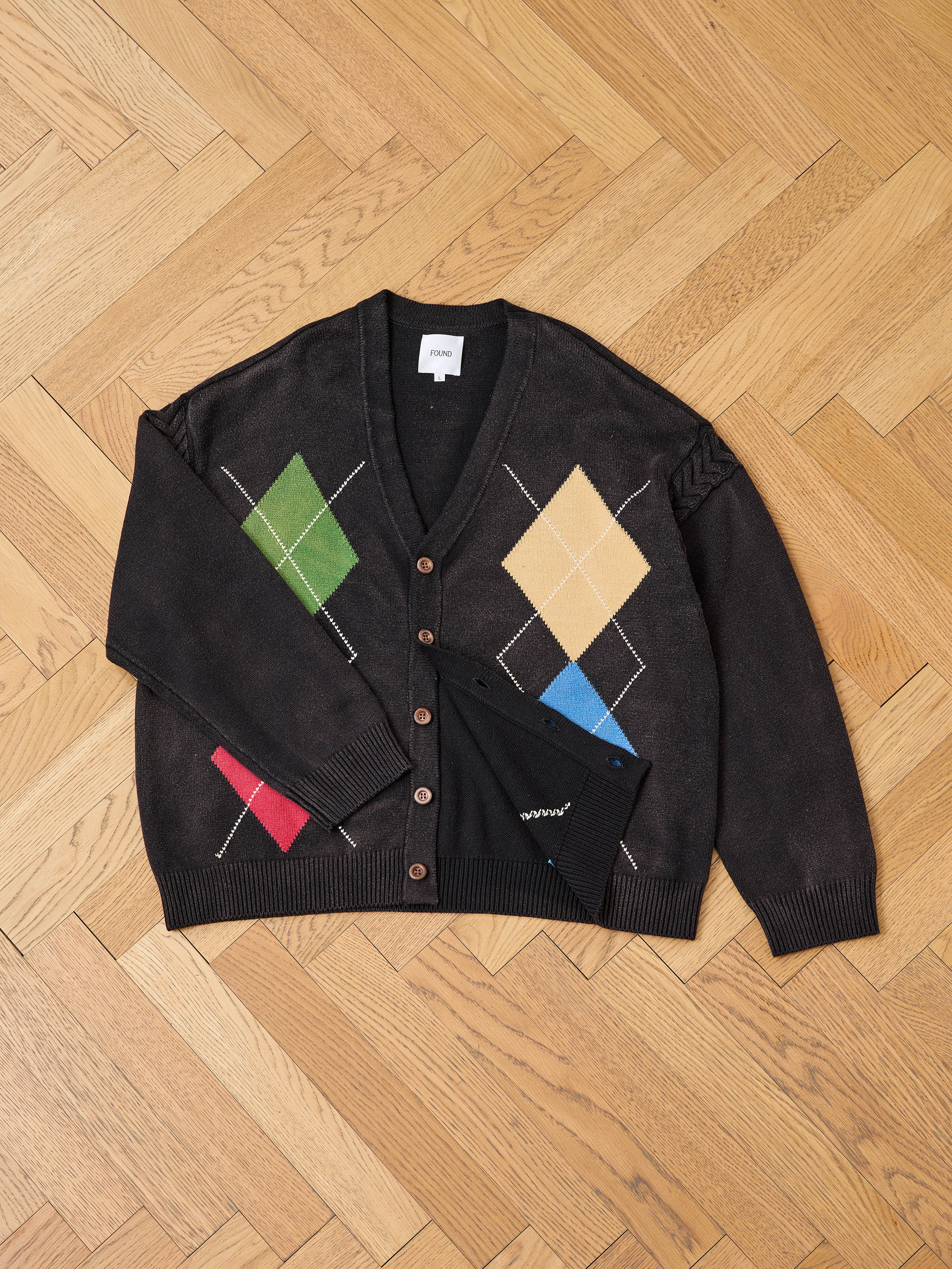A dark brown unisex FOUND Argyle Knit Cardigan with a multicolored pattern, button front, and collar label is laid flat on a wooden herringbone floor.
