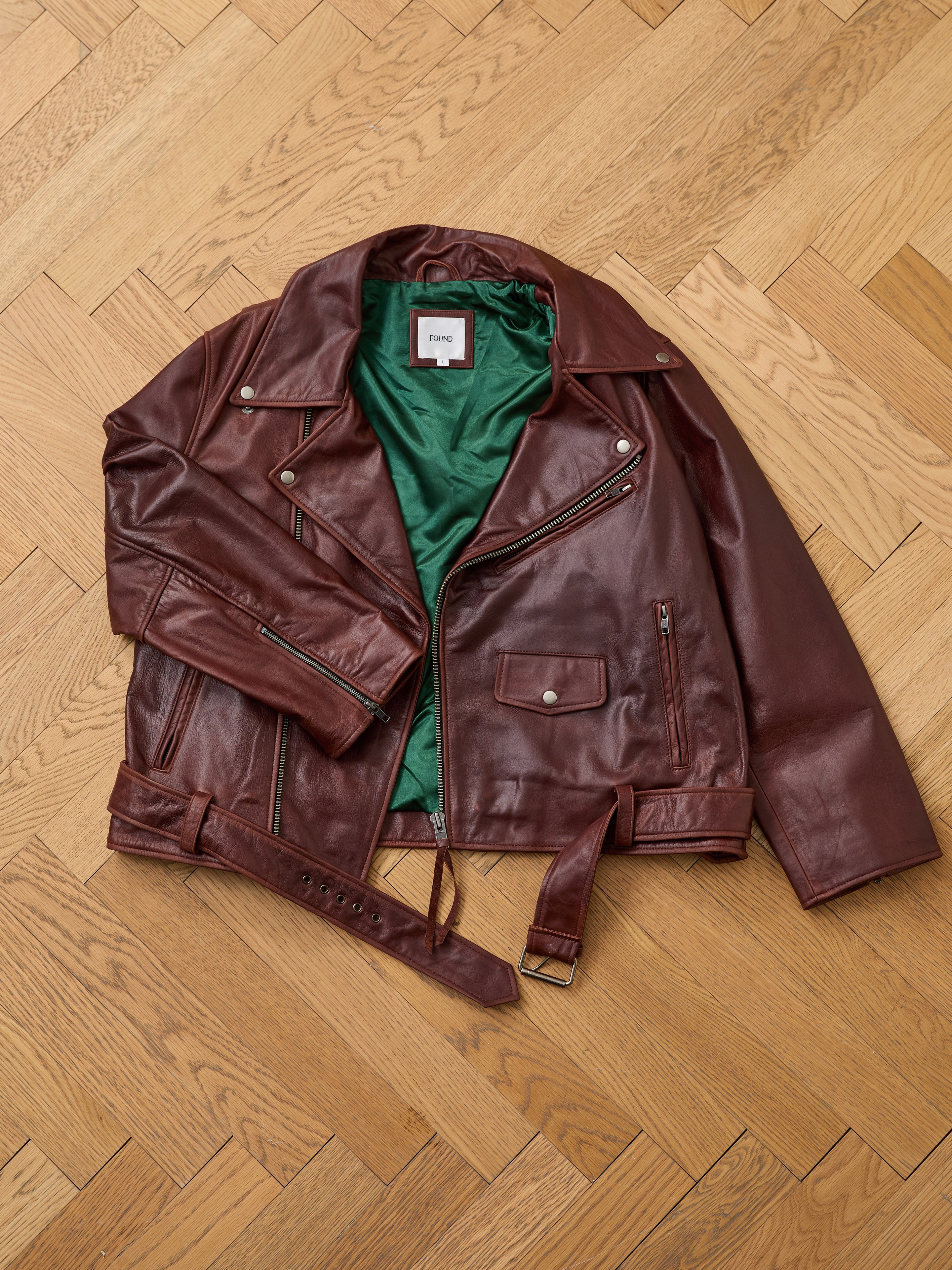 The FOUND Leather Biker Jacket, a unisex design in brown distressed leather with silver zippers and a green inner lining, is displayed flat on a wooden parquet floor.
