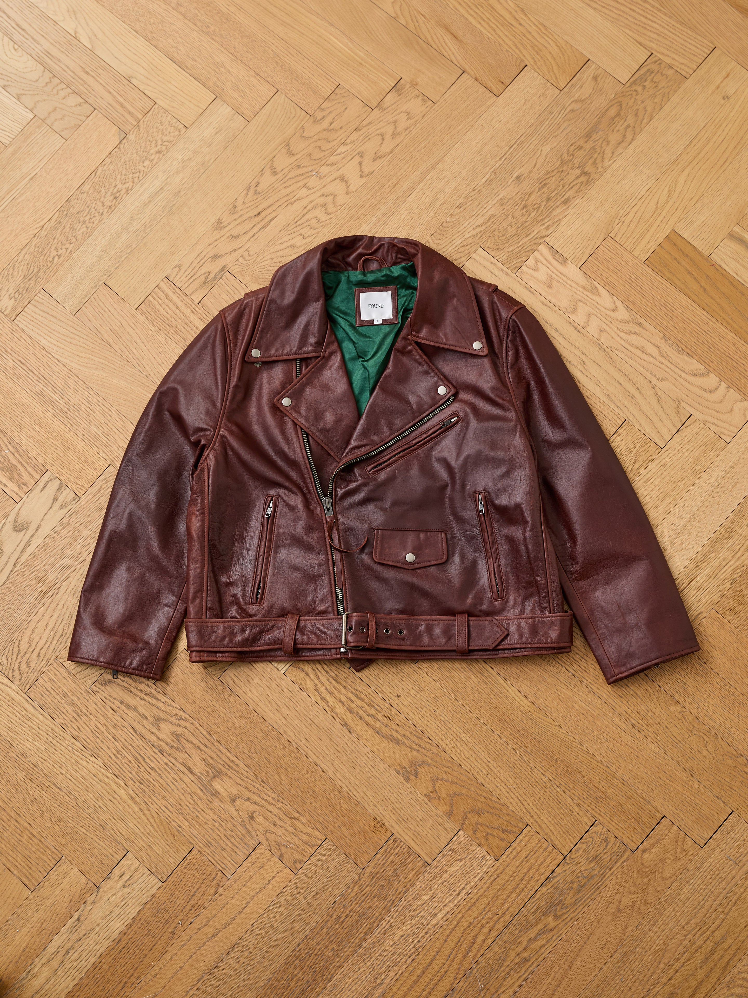 The FOUND Leather Biker Jacket in brown, featuring green lining, silver zippers, and a belt, is displayed flat on a wooden herringbone floor.