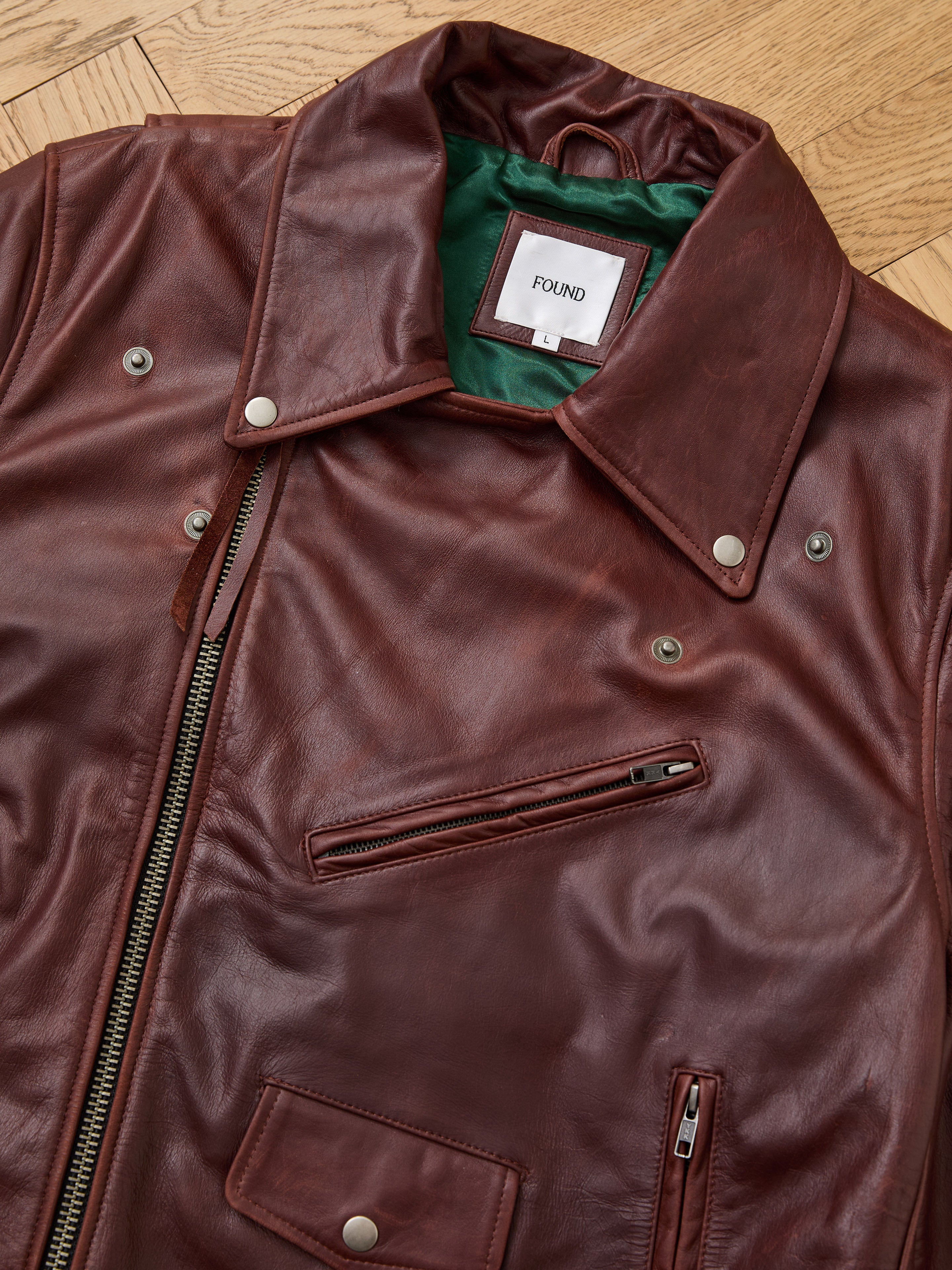 The Leather Biker Jacket by FOUND features brown distressed leather, a zippered front, metal snaps, multiple pockets, a green interior lining, and the signature "FOUND" label—shown laid flat on a wooden floor.