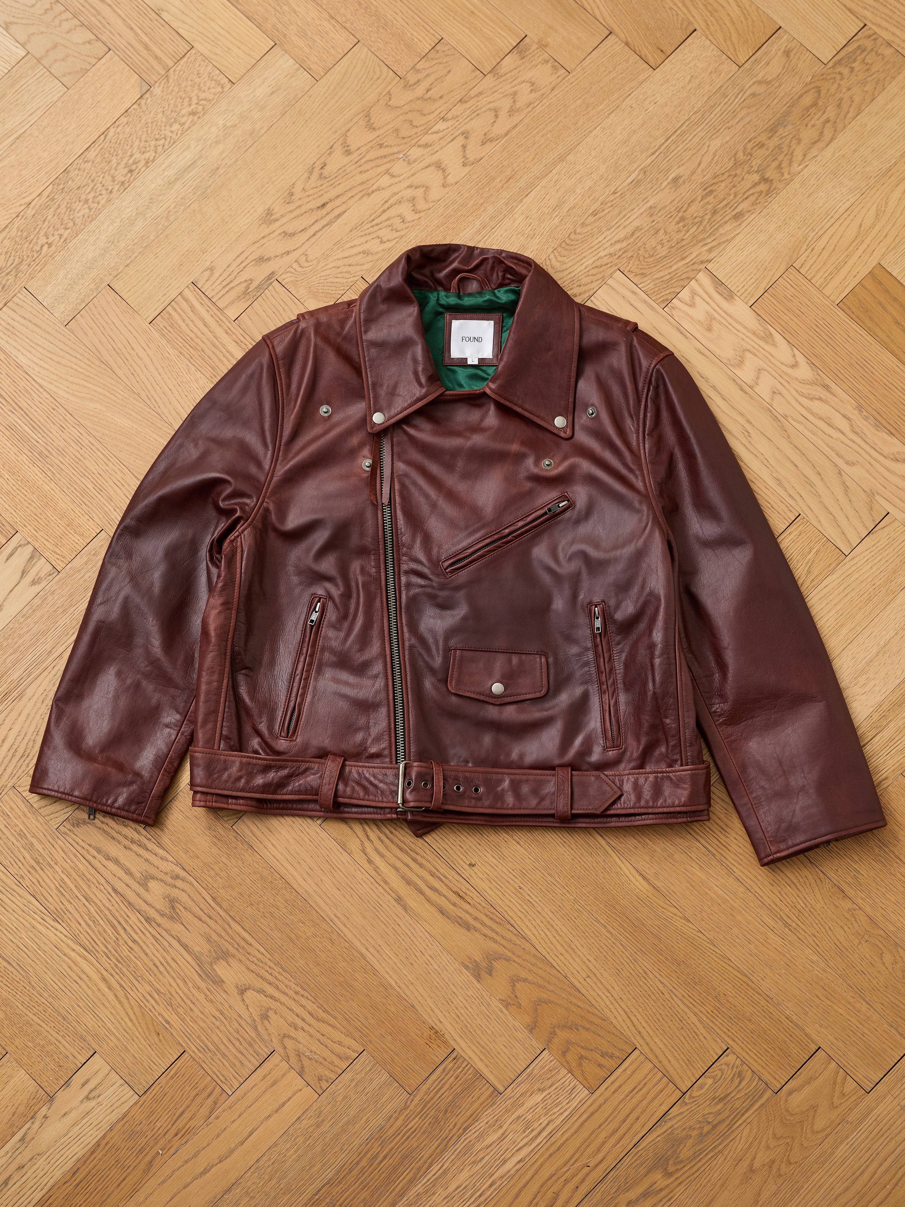 The FOUND Leather Biker Jacket, a brown unisex leather jacket with multiple zippered pockets and a belt, is laid flat on a light wood herringbone floor.