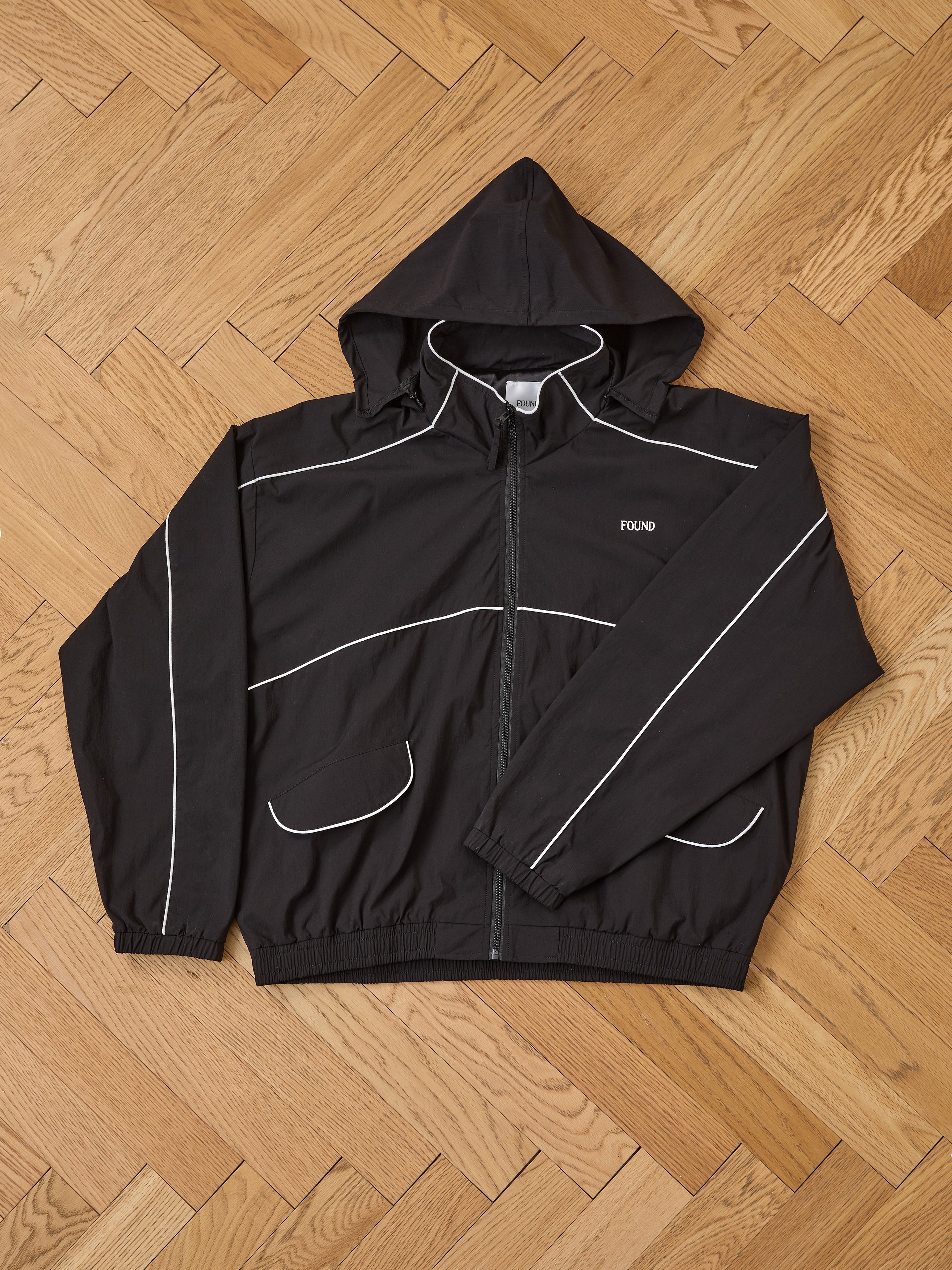 The FOUND Arena Stripe Track Jacket is a unisex black jacket made from lightweight technical fabric, with white piping, a front zipper, two flap pockets, and "FOUND" on the chest. It’s shown laid flat on a wooden parquet floor.