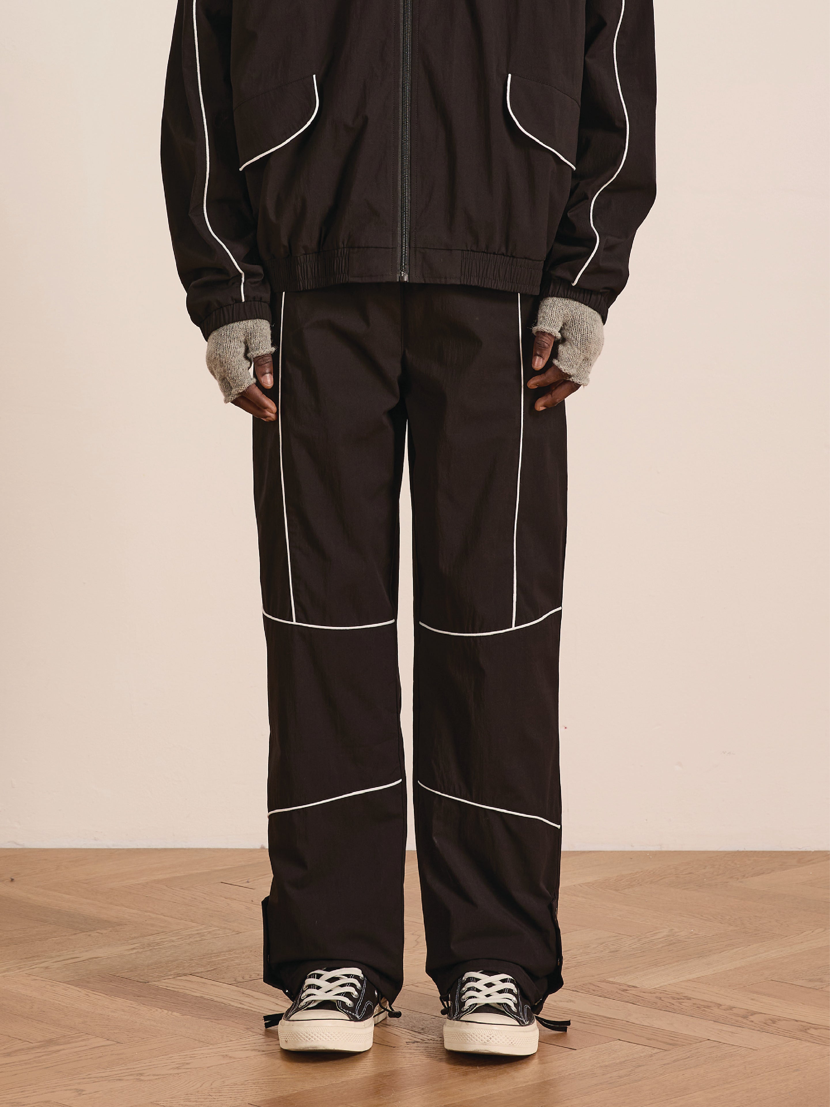 A person stands on a wooden floor wearing Found's Arena Stripe Track Pants—black with white piping and an adjustable waistband—paired with a black zip-up jacket, gray fingerless gloves, and black sneakers for a unisex sportswear look.