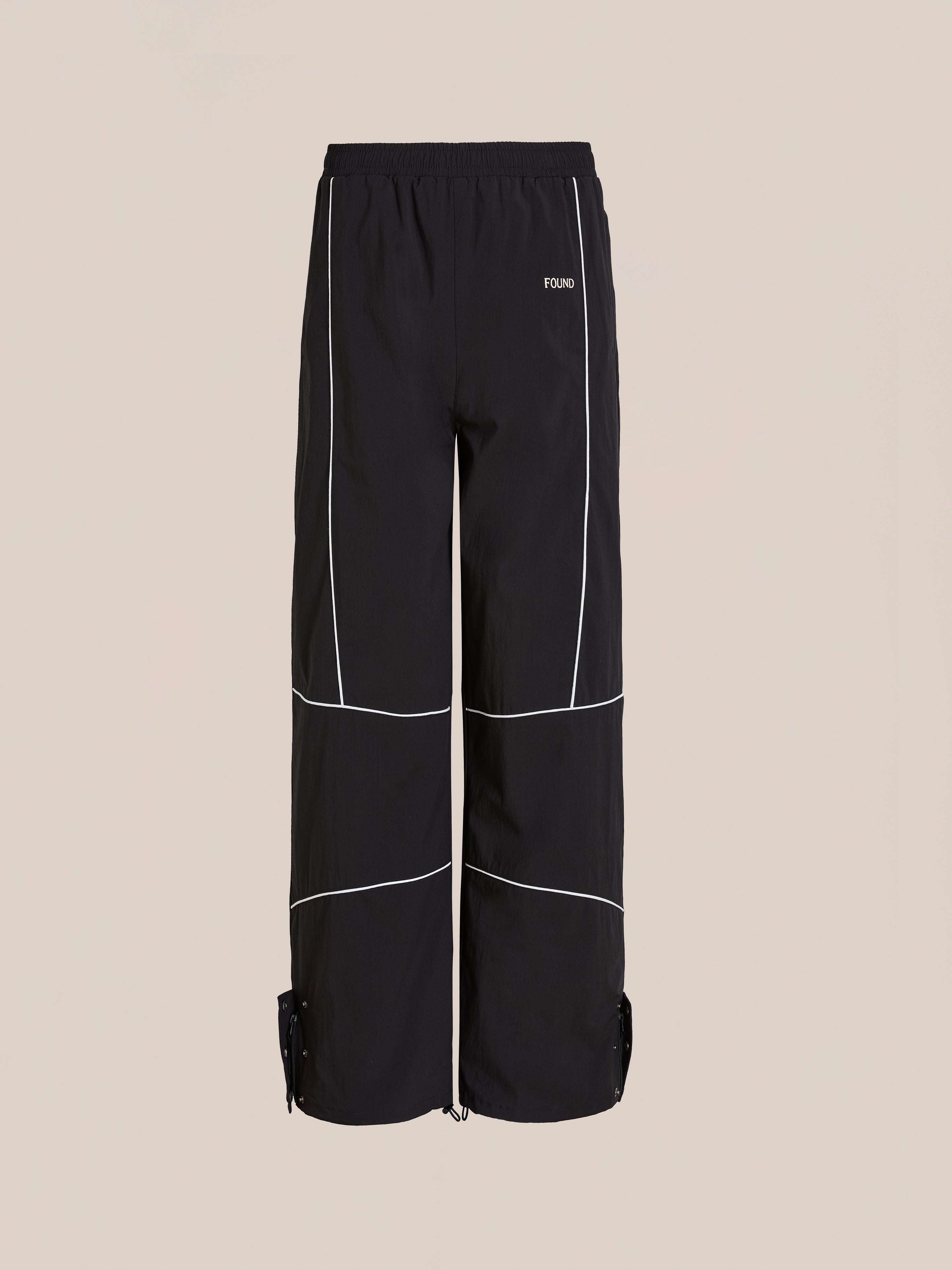 The Found Arena Stripe Track Pants feature a black relaxed fit, white piping, adjustable waistband and ankle tabs, and a subtle left text logo. Shown on a beige background—versatile unisex sportswear for any occasion.