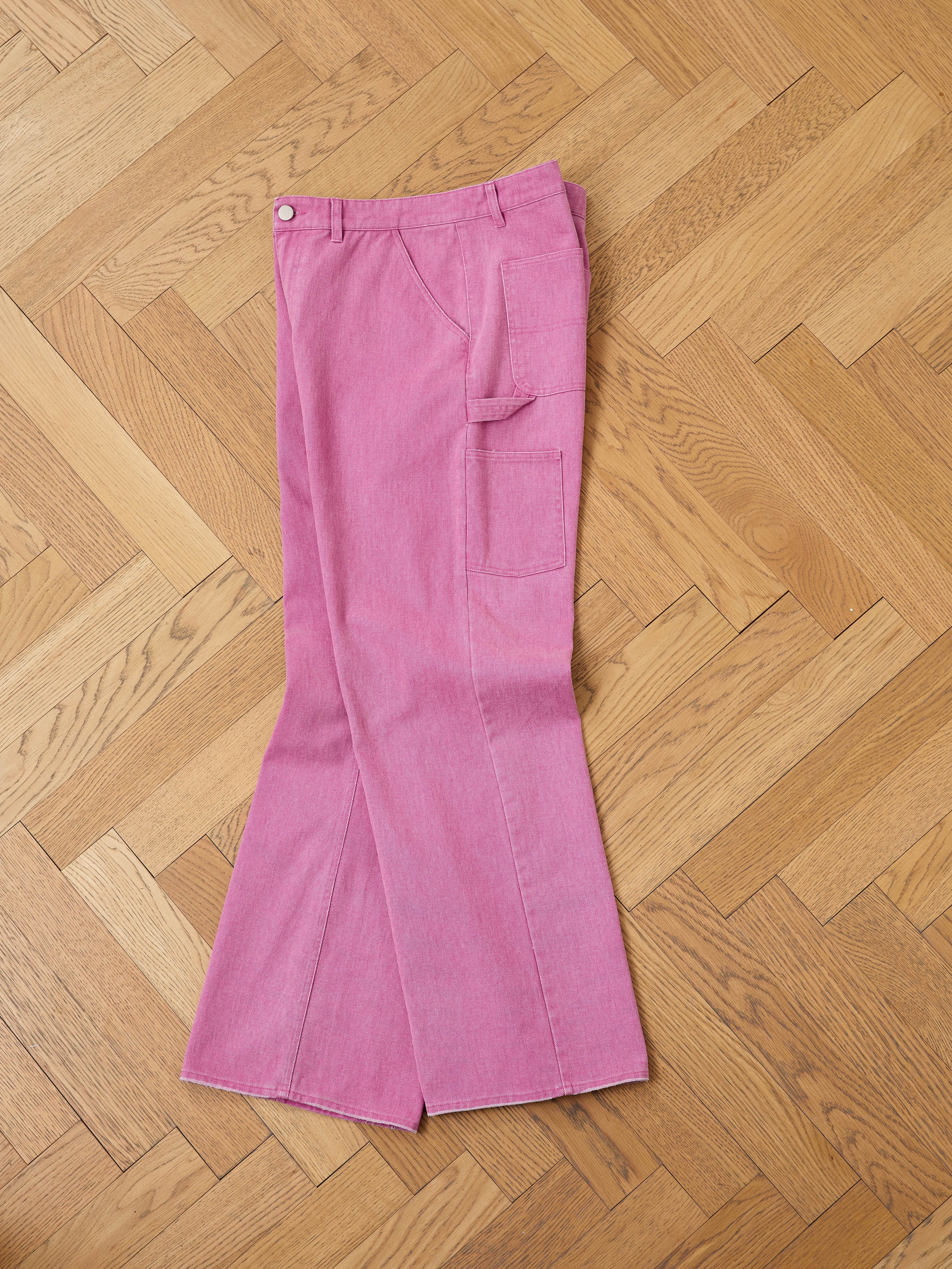 FOUND Flare Work Pants in pink denim with flared legs and a side pocket are laid flat on a light brown herringbone floor, offering stylish unisex workwear.
