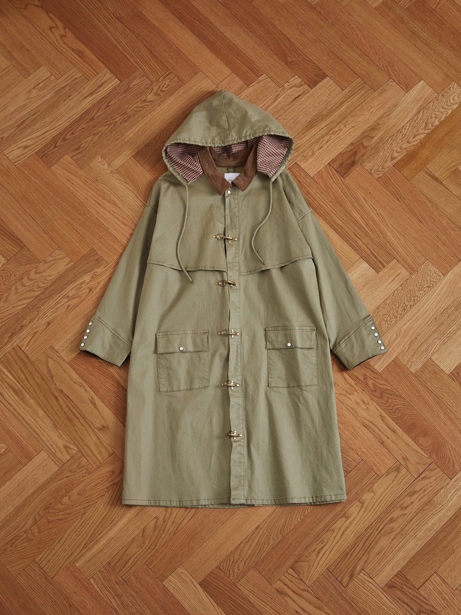 A green Prairie Buckle Coat made of waxed cotton lays on a wooden floor, showcasing its water resistance by Found.
